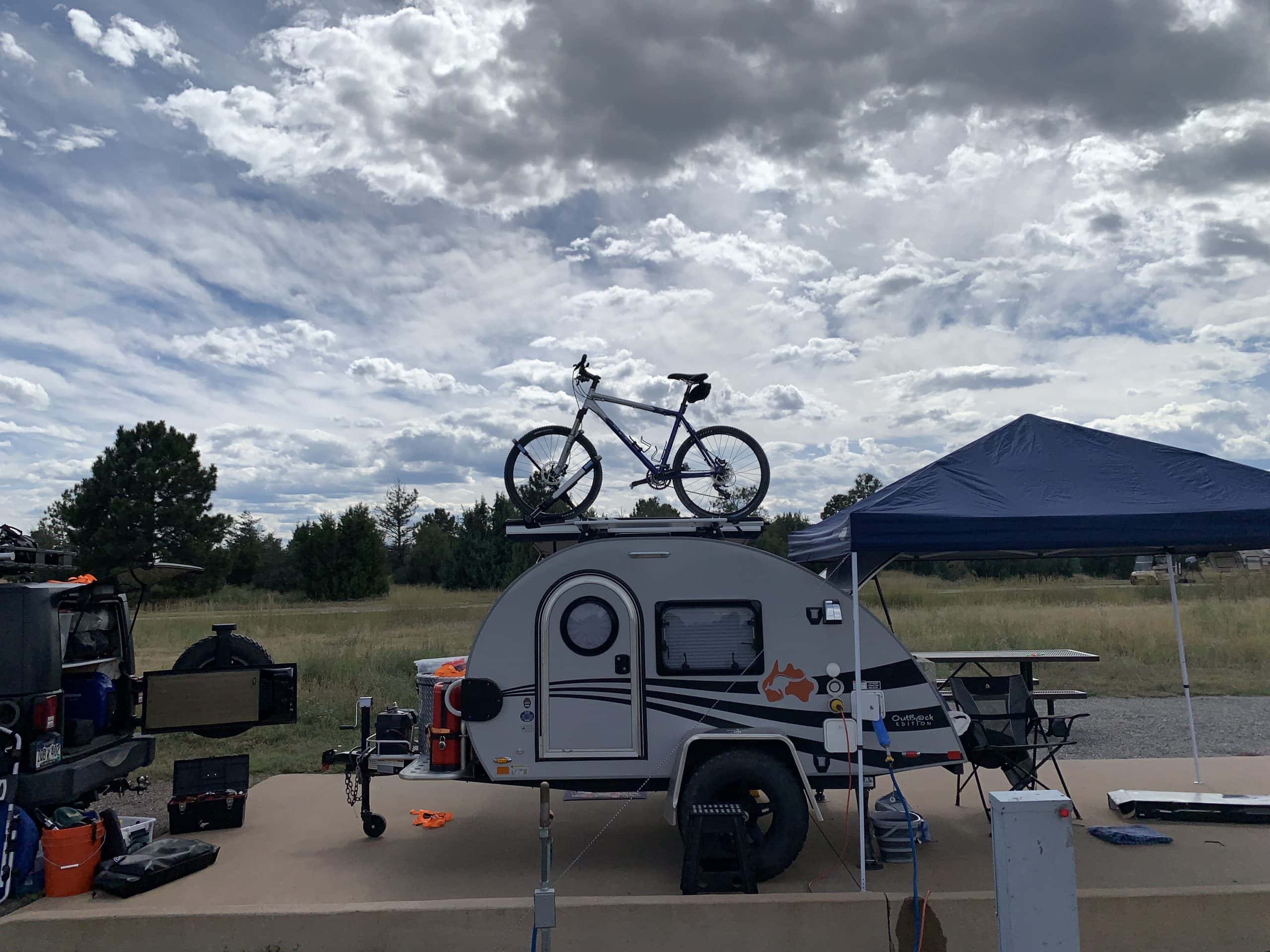A small teardrop camper with a bicycle mounted on its roof is parked beside a blue canopy tent. Camping gear, a table, and chairs are set up nearby. Cloudy sky and grassy fields are in the background, with trees lining the horizon.