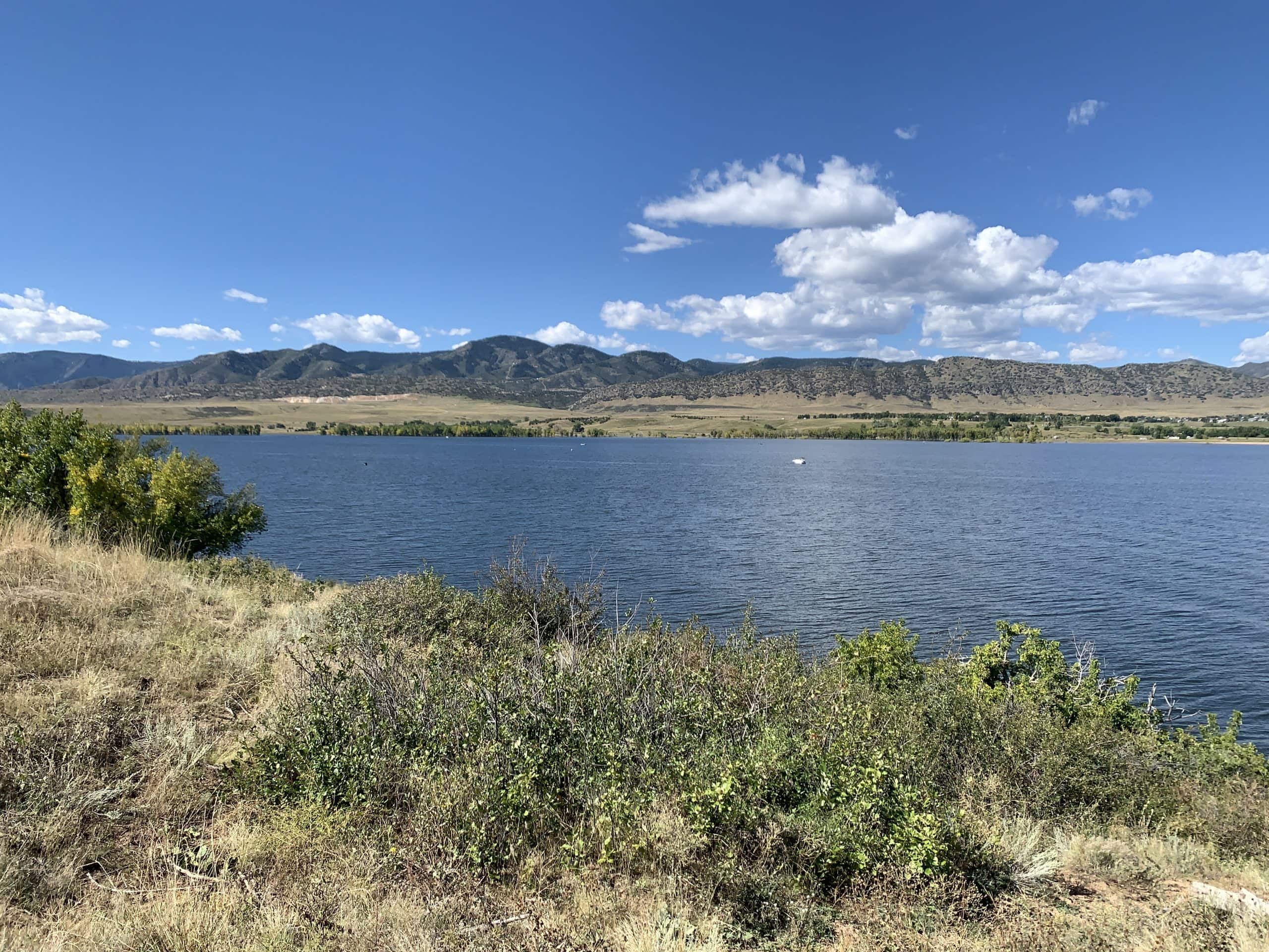 A calm blue lake stretches across the foreground, bordered by dry grass and green shrubs. In the background, low, rugged mountains rise beneath a mostly clear blue sky with some scattered white clouds.