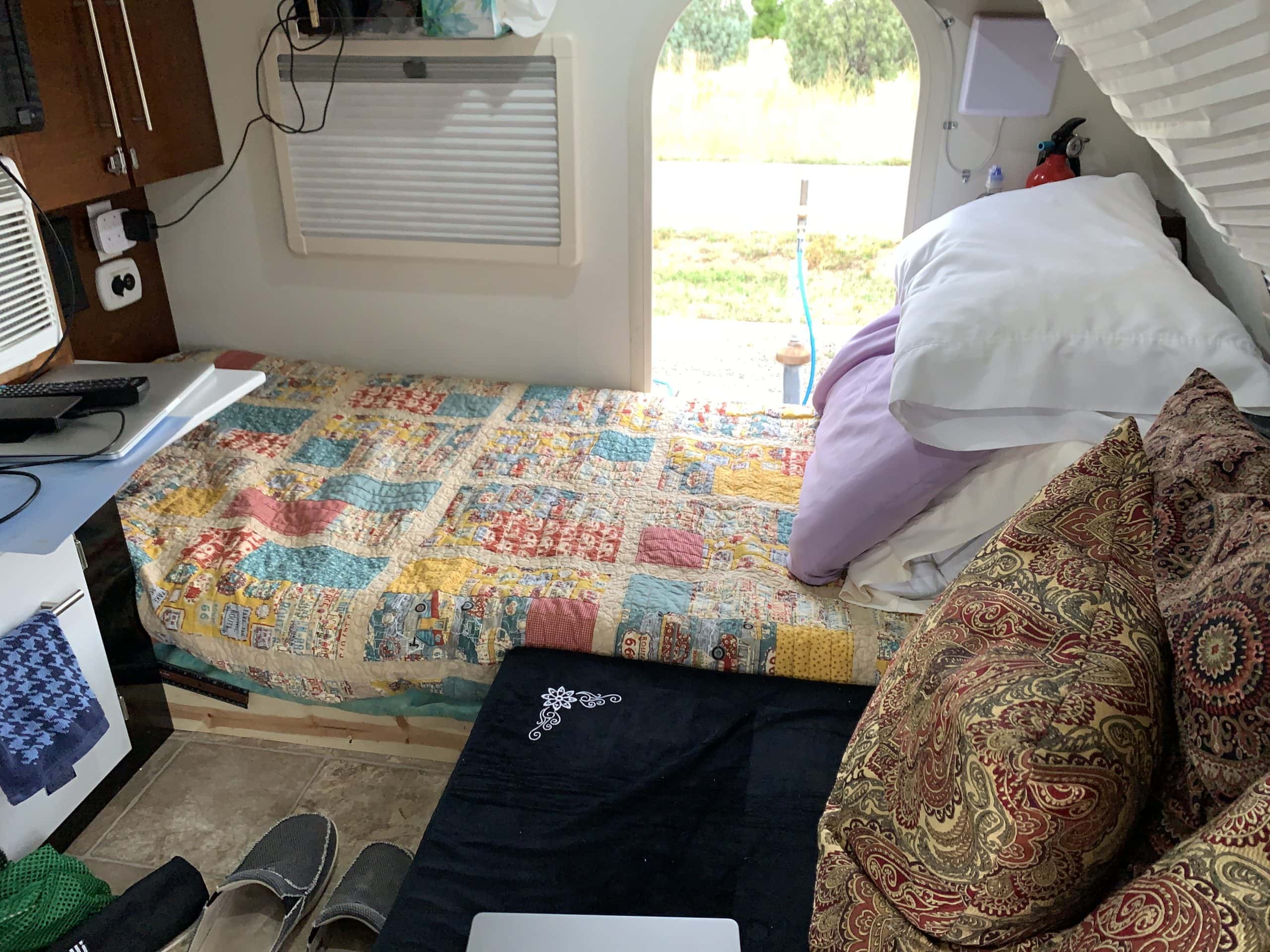 A cozy RV interior with a colorful patchwork quilt on a bed, stacked pillows, patterned cushions, a small window with a shade, a guitar, slippers, a laptop, and a blue-and-black towel. Sunlight streams in from the open door, revealing greenery outside.