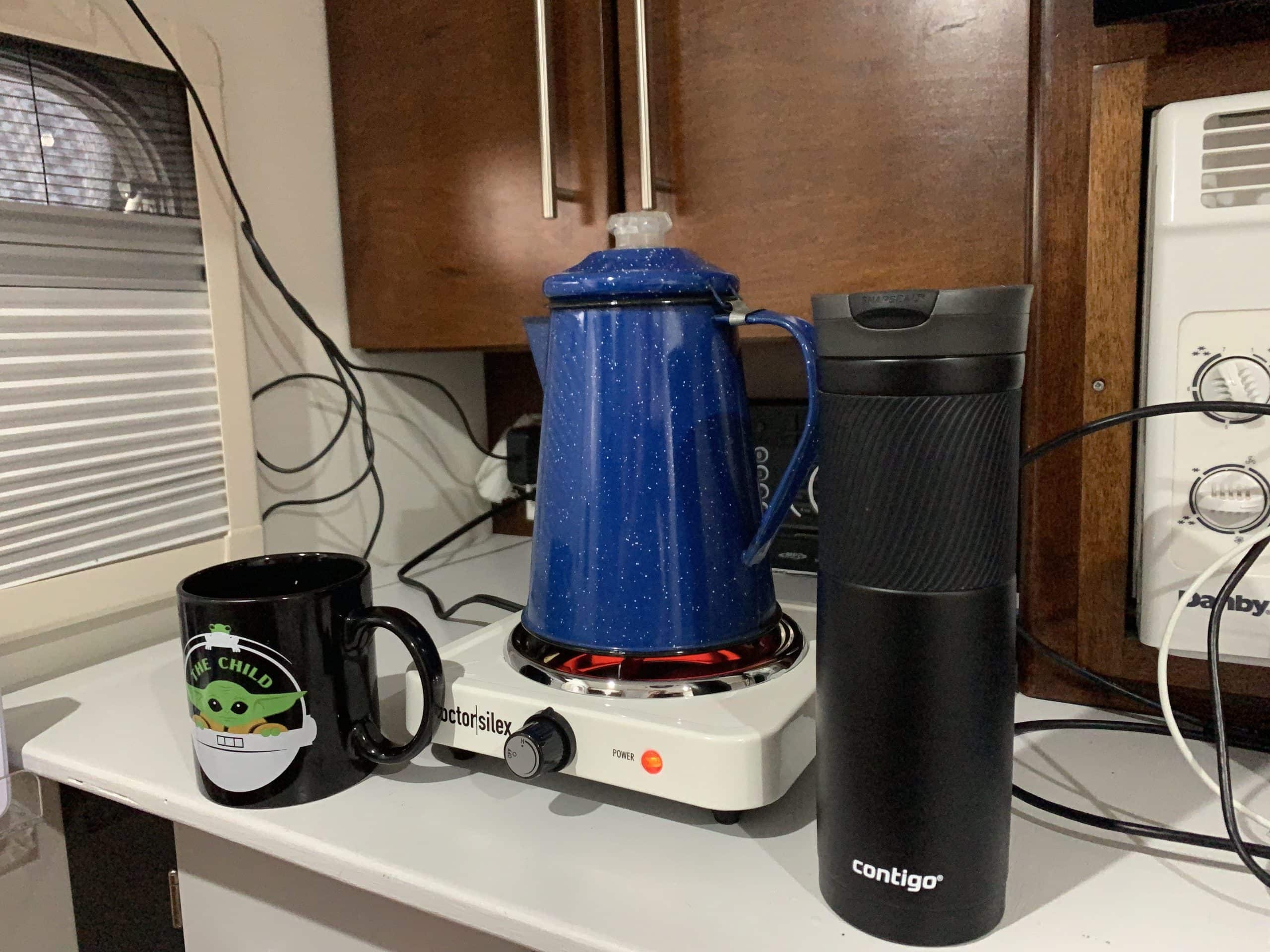A blue speckled percolator sits on a white electric hot plate next to a black Contigo travel mug and a black mug with “The Child” and a cartoon of Baby Yoda. The scene is set on a white countertop in a cozy kitchen with wooden cabinets and appliances.
