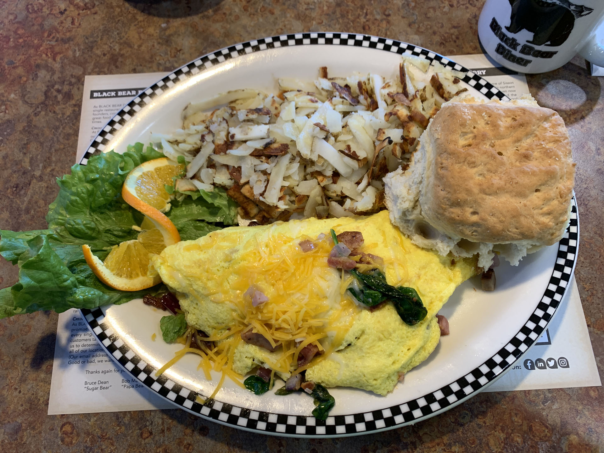 A hearty omelet with shredded cheese, spinach, and ham sits beside crispy hash browns, a biscuit, lettuce, and orange slices—all served on a checkered-rim plate at Black Bear Diner, capturing the classic American diner experience.