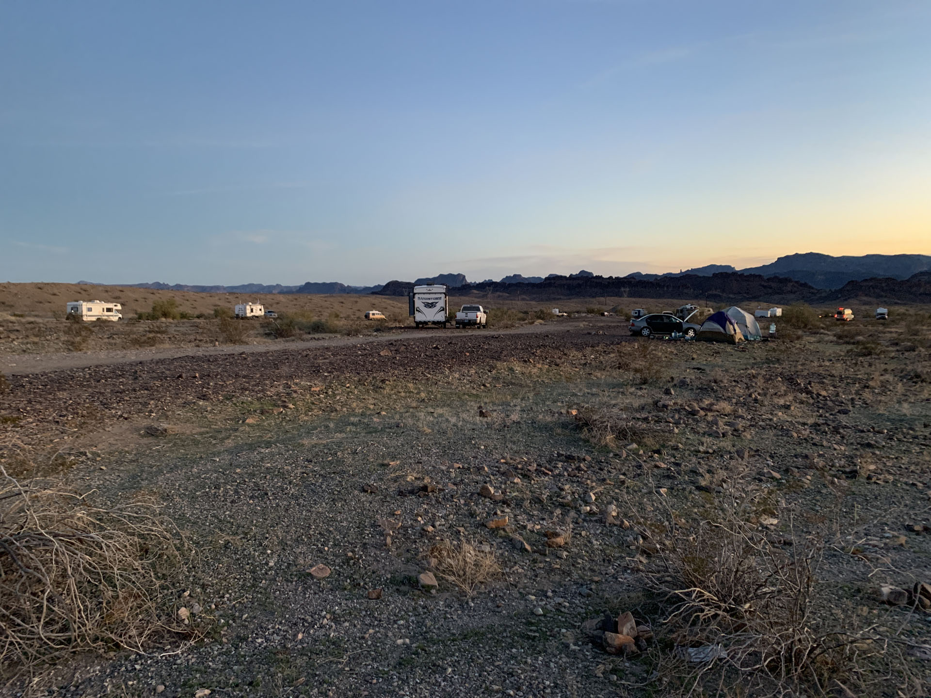 Campsite in a rocky desert at sunset near Havasu, with scattered RVs, cars, and tents along a dirt road—classic BLM dispersed camping. Low brush and rocks cover the ground; distant hills frame this nominal place to sleep under a clear sky.