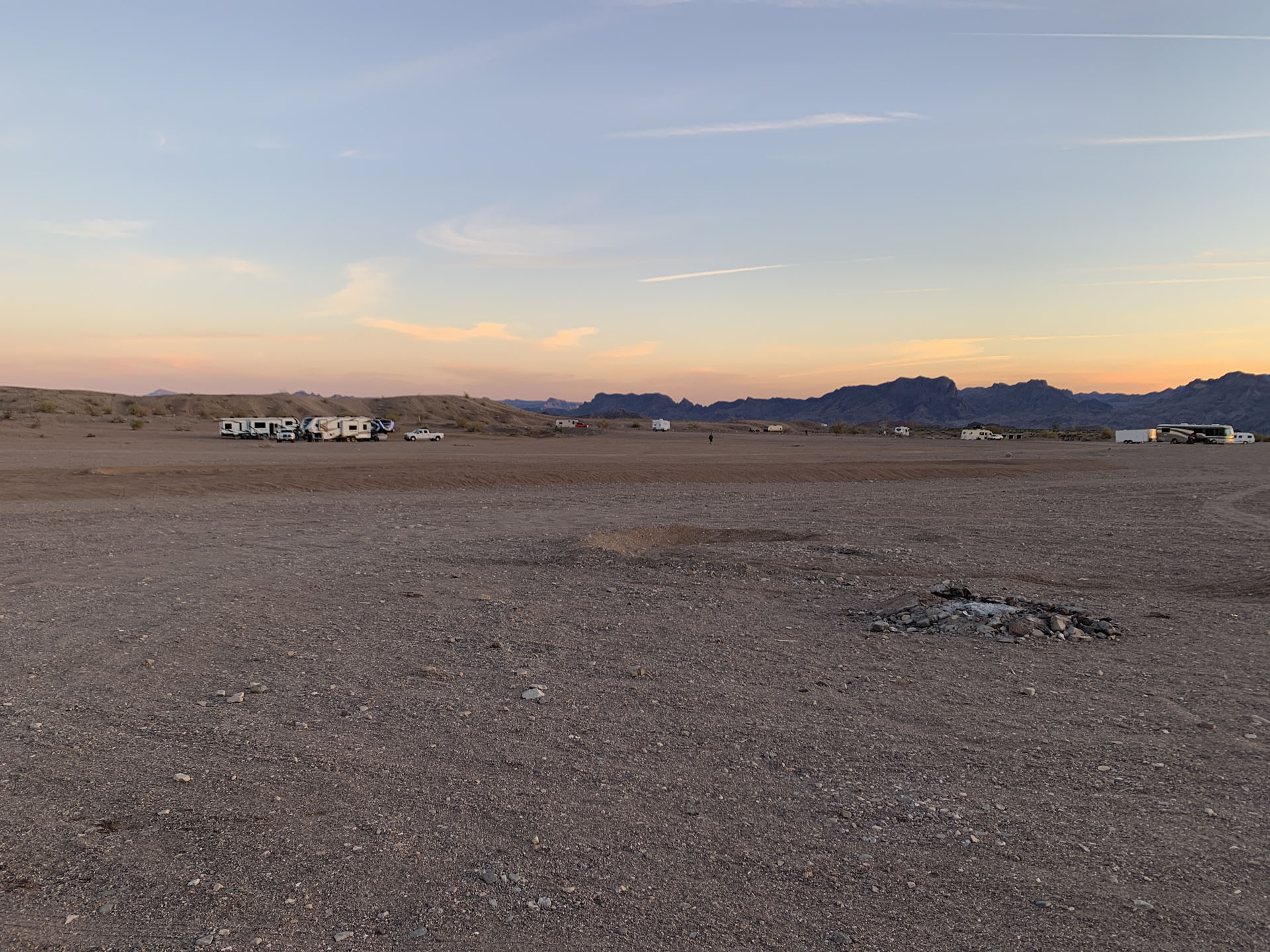 A wide, barren BLM Dispersed desert landscape at sunset with a few RVs parked in the distance. The sky is pastel blue and orange, mountains line the horizon, and a small fire pit marks this nominal place to sleep in Havasu’s dry, rocky terrain.