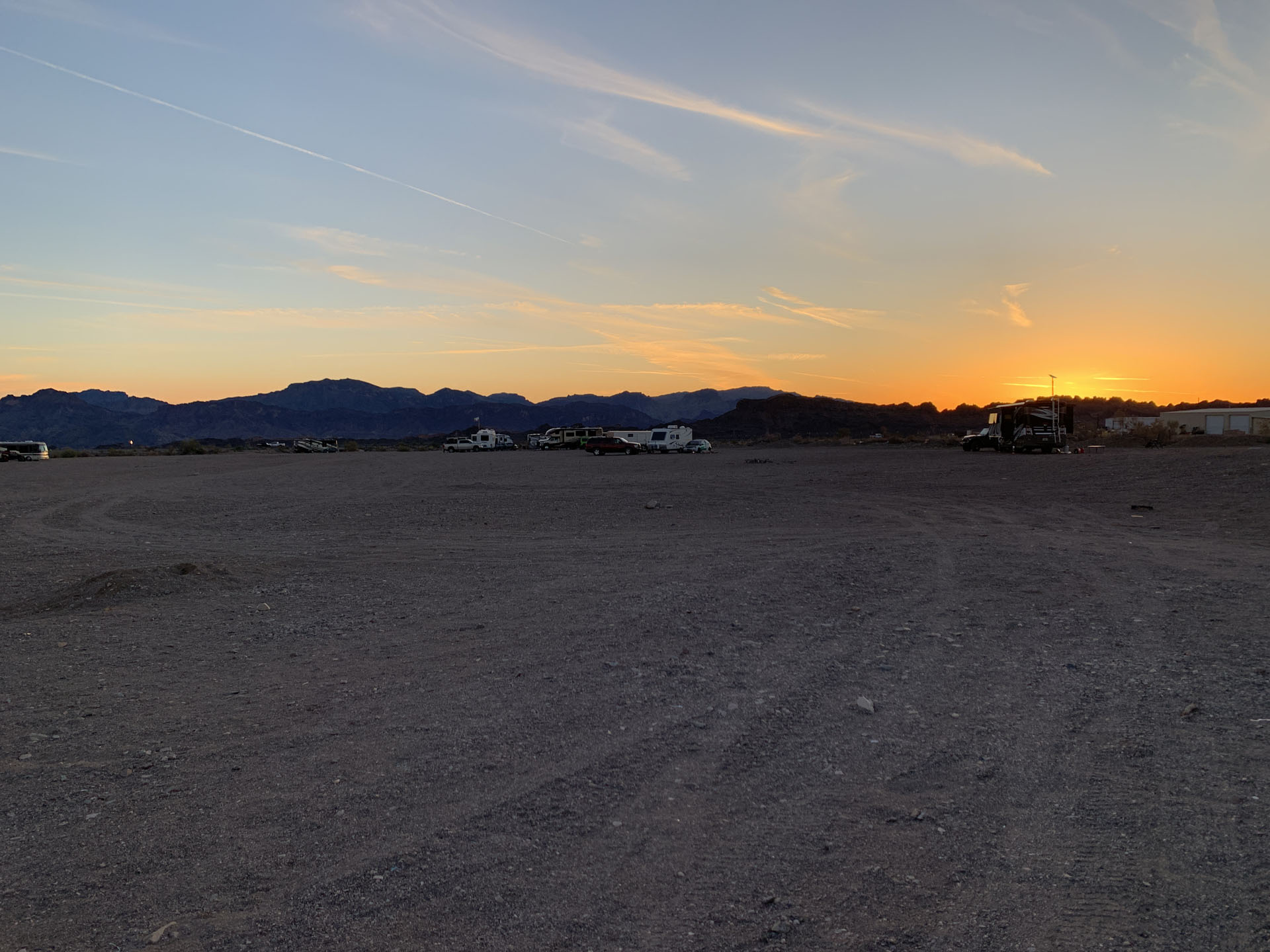 A wide, gravelly desert landscape at sunset with distant mountains against an orange and blue sky. Several RVs are parked near the horizon, offering a place to sleep at Havasu BLM Dispersed, while contrails streak diagonally across the sky.