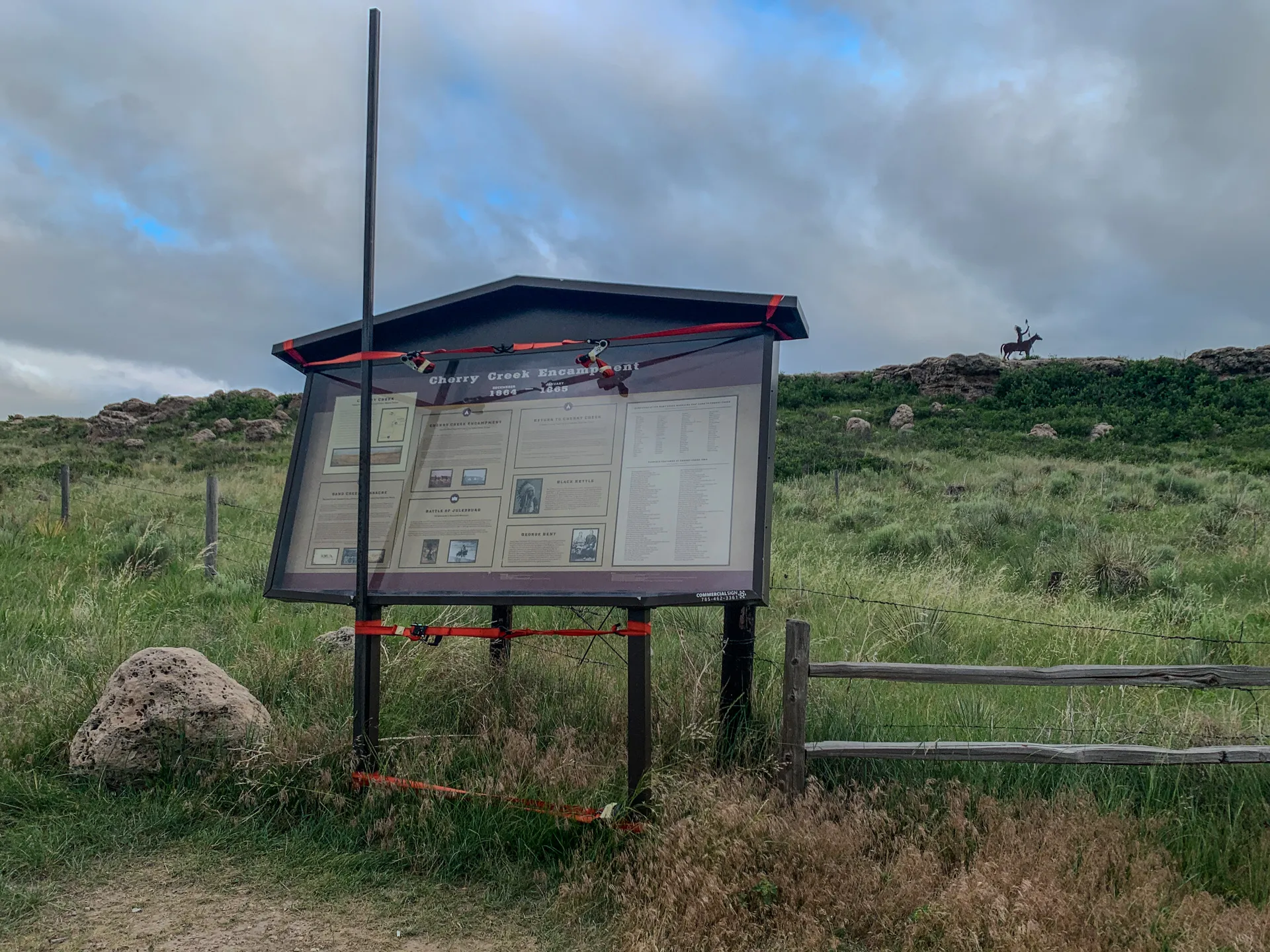 An outdoor informational sign stands in a grassy field under a cloudy sky, providing details about "Cherry Creek Encampment" with a fenced area and rocky hill in the background.