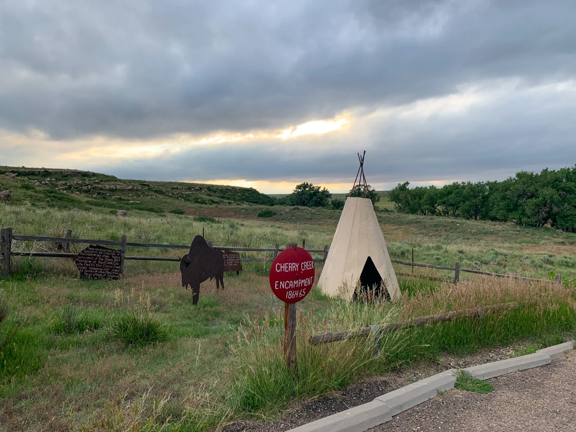 A teepee and a metal buffalo sculpture are displayed in a grassy area with a wooden fence and a sign reading "Cherry Creek Encampment 1864-65." A cloudy sky is visible in the background.