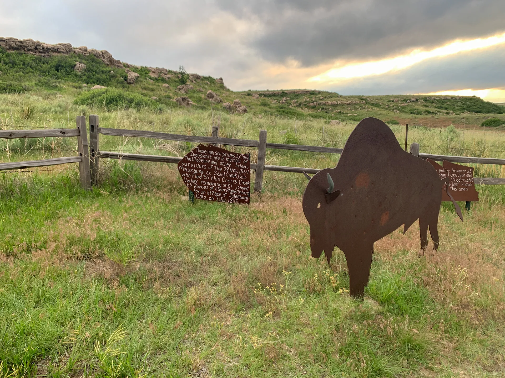 A grassy field with a wooden fence displaying a metal bison silhouette and an informational arrow-shaped sign. Cloudy sky and rolling hills in the background.