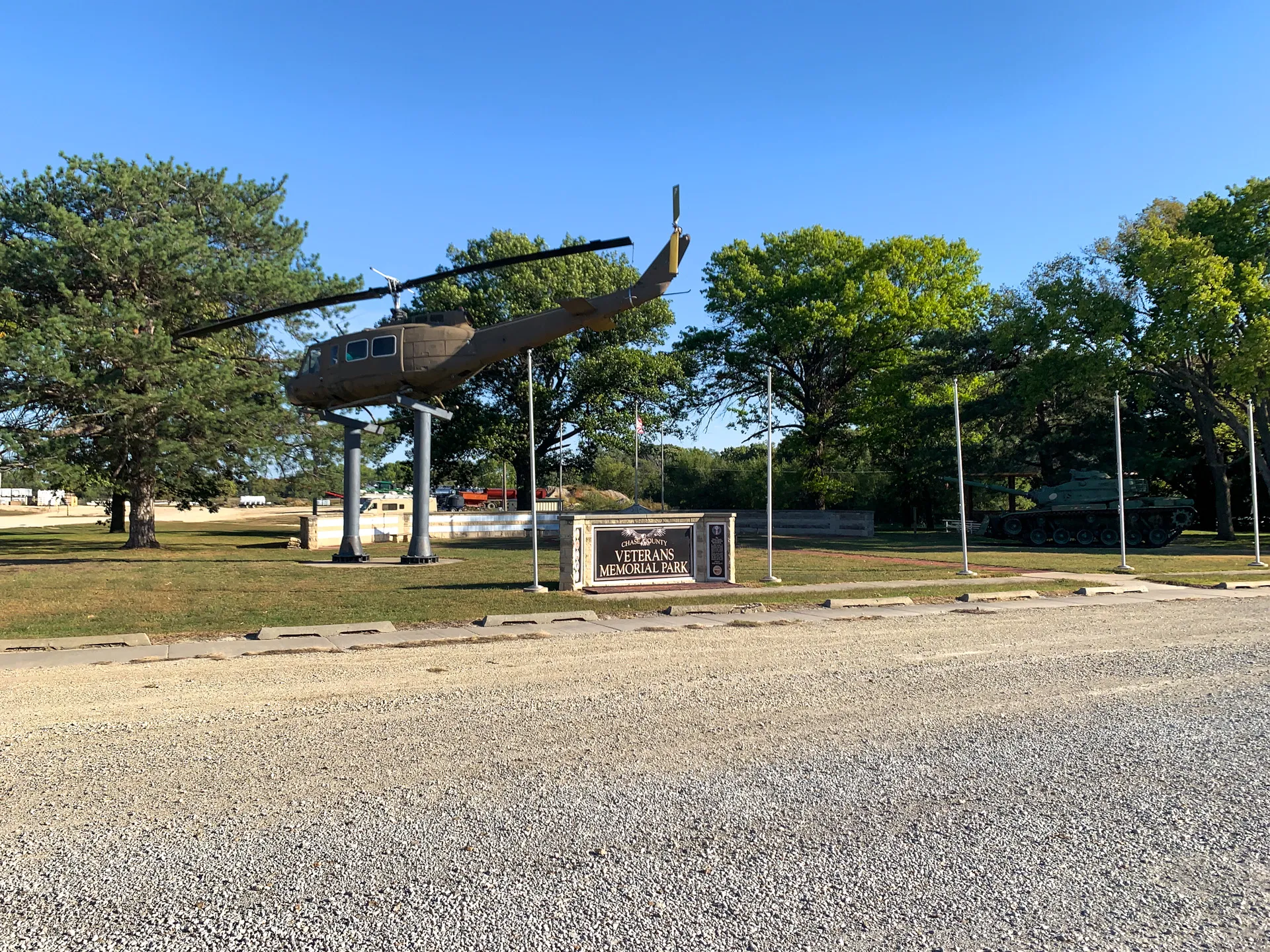 A helicopter mounted on stands and a tank displayed Chase County Veterans Memorial Park, surrounded by trees and blue sky.