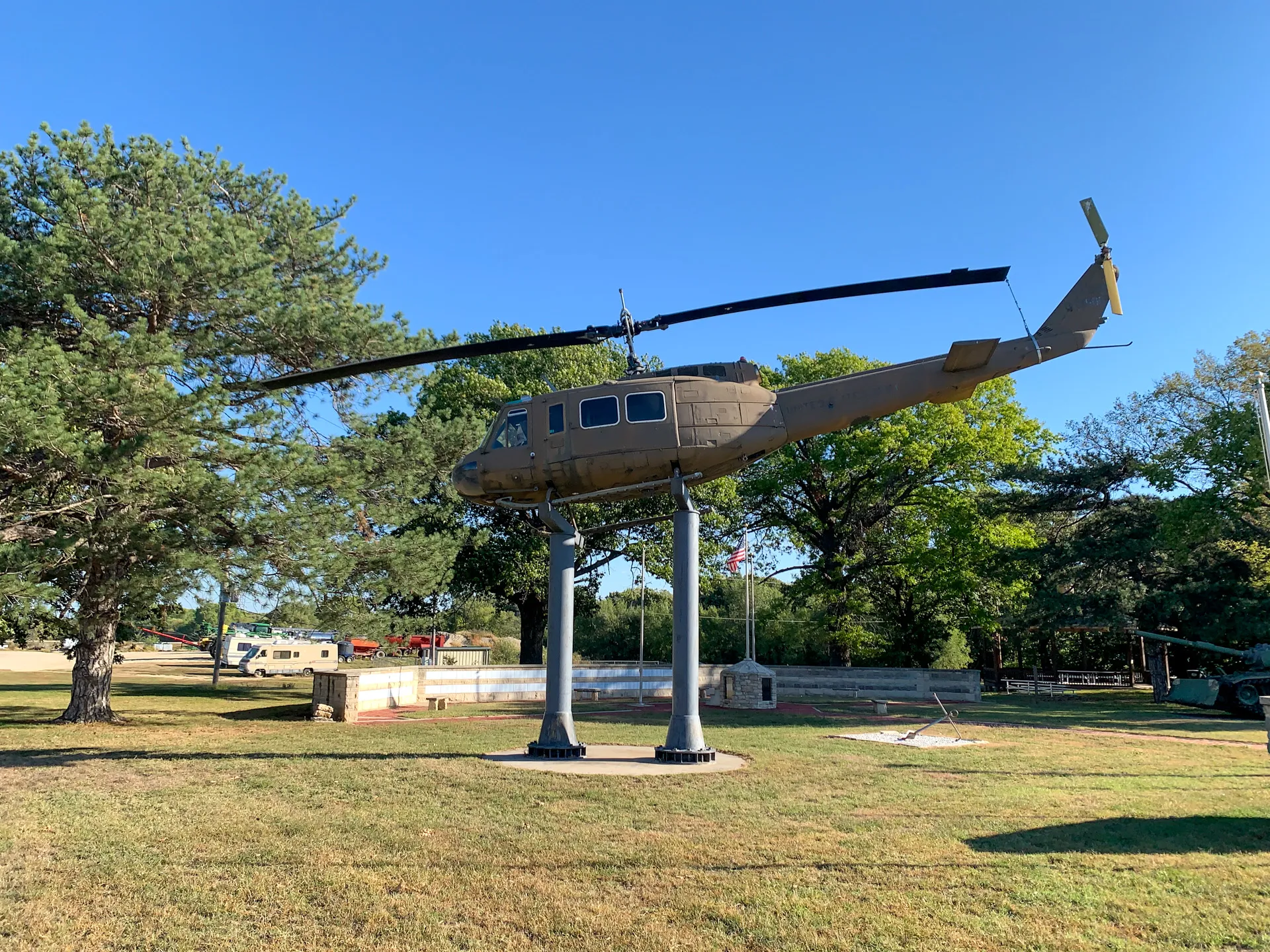 A military helicopter is mounted on four poles in a grassy park area with trees and vehicles in the background.