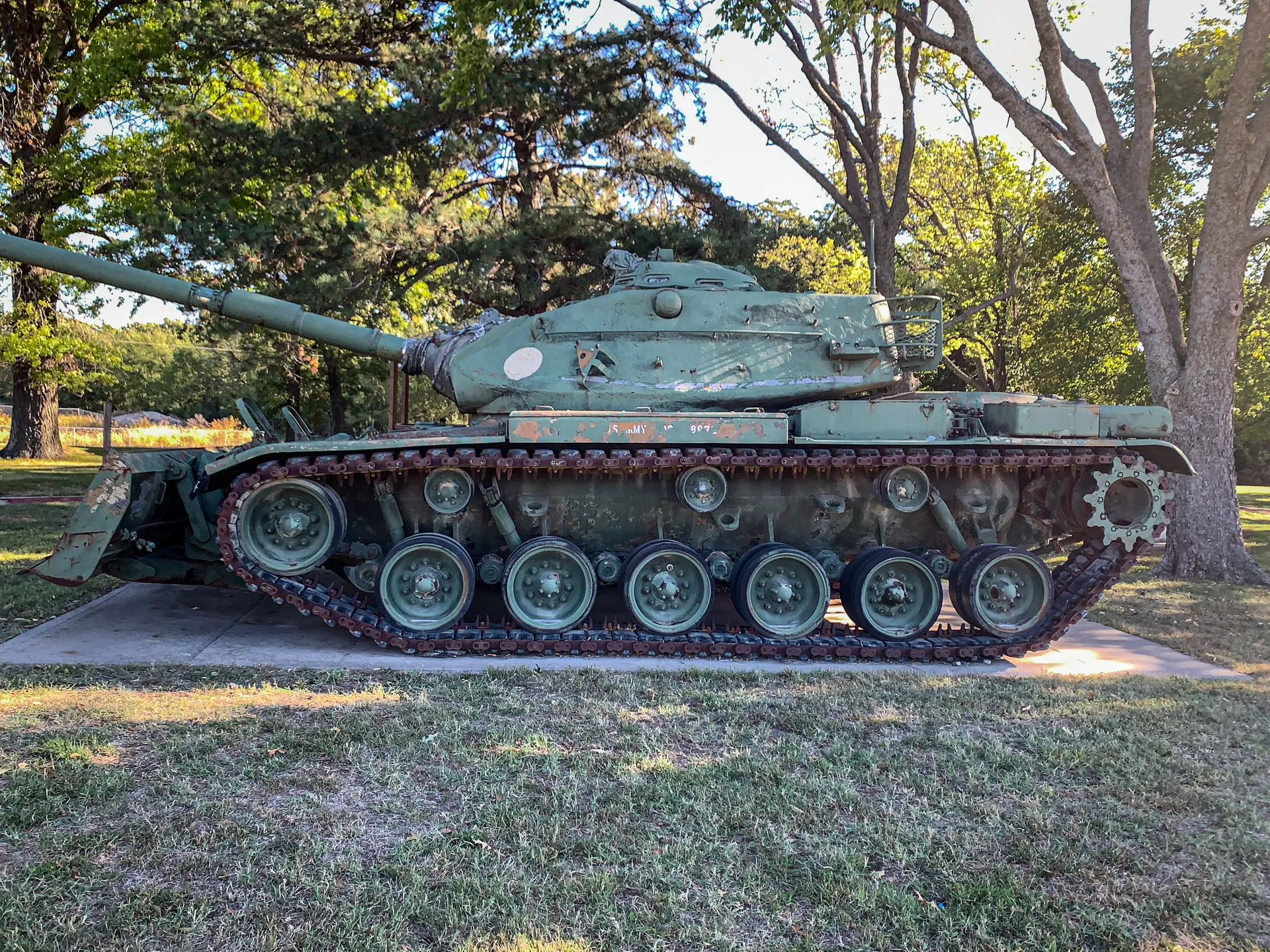 A green military tank with a large cannon on a concrete platform in a park, surrounded by trees and grass.