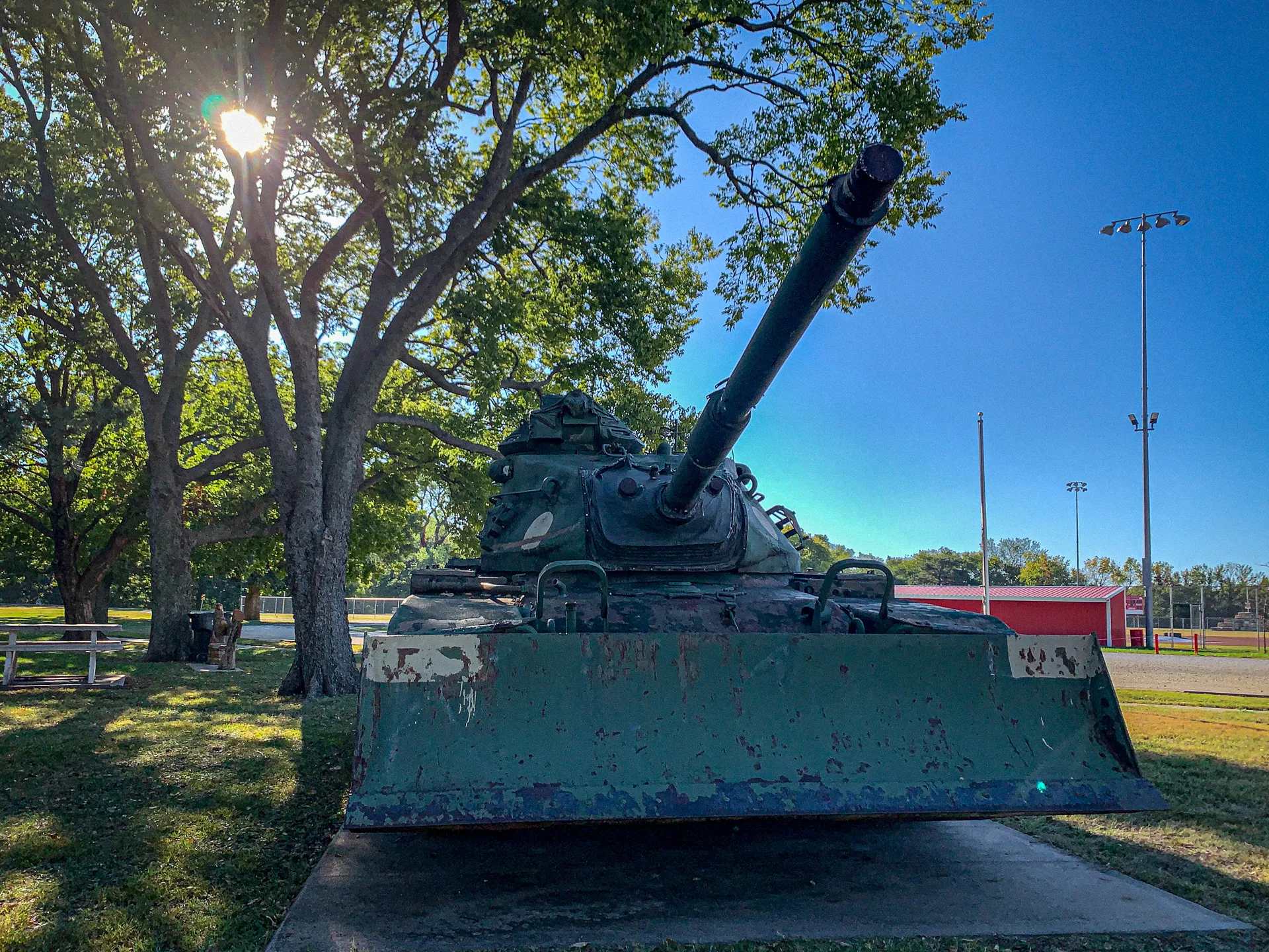 A weathered, decommissioned tank displayed outdoors on a concrete slab, surrounded by trees. A sports field with poles and backboards is visible in the background.