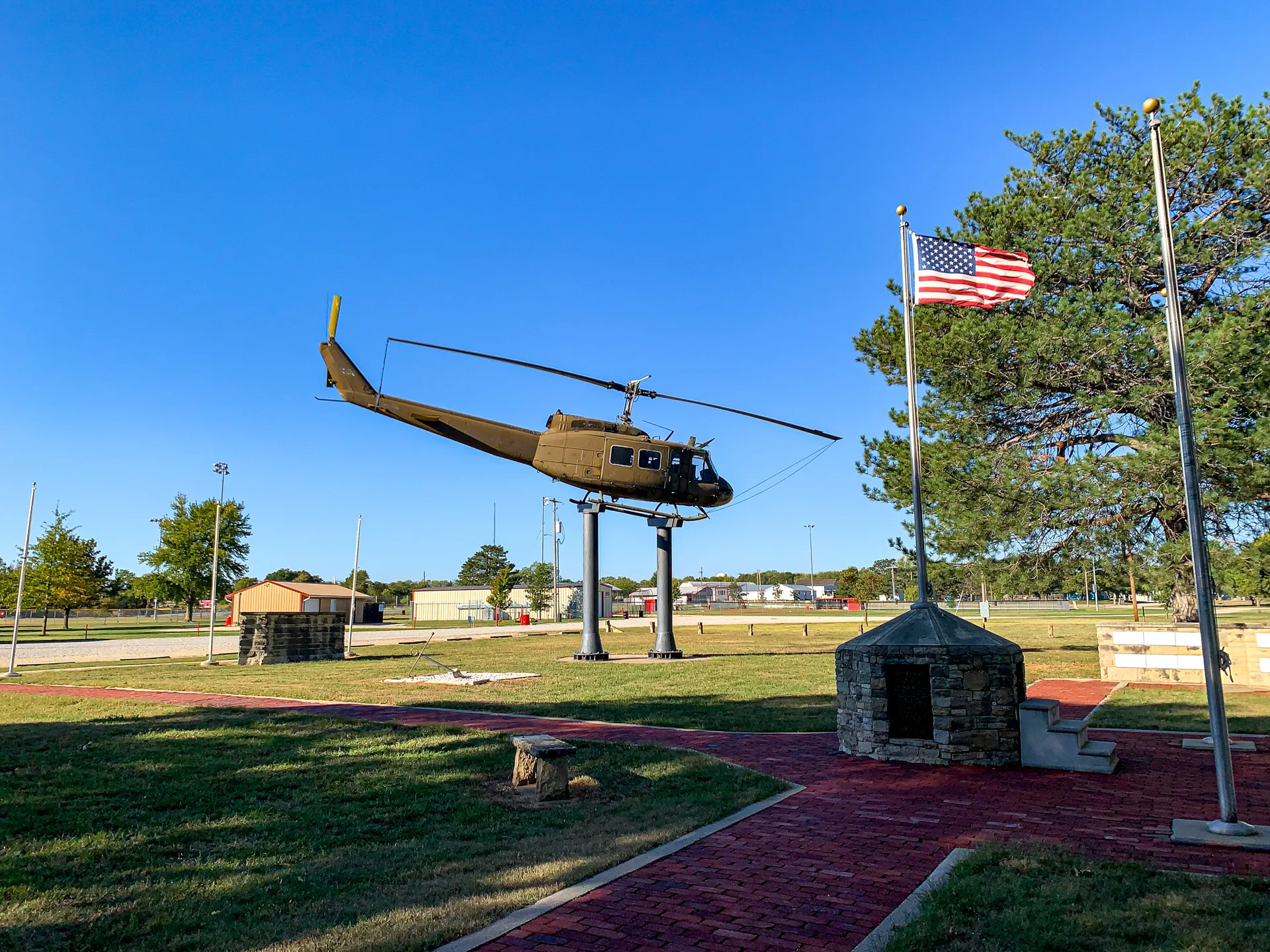 A military helicopter is displayed on a stand in an open park area with an American flag on a flagpole nearby.