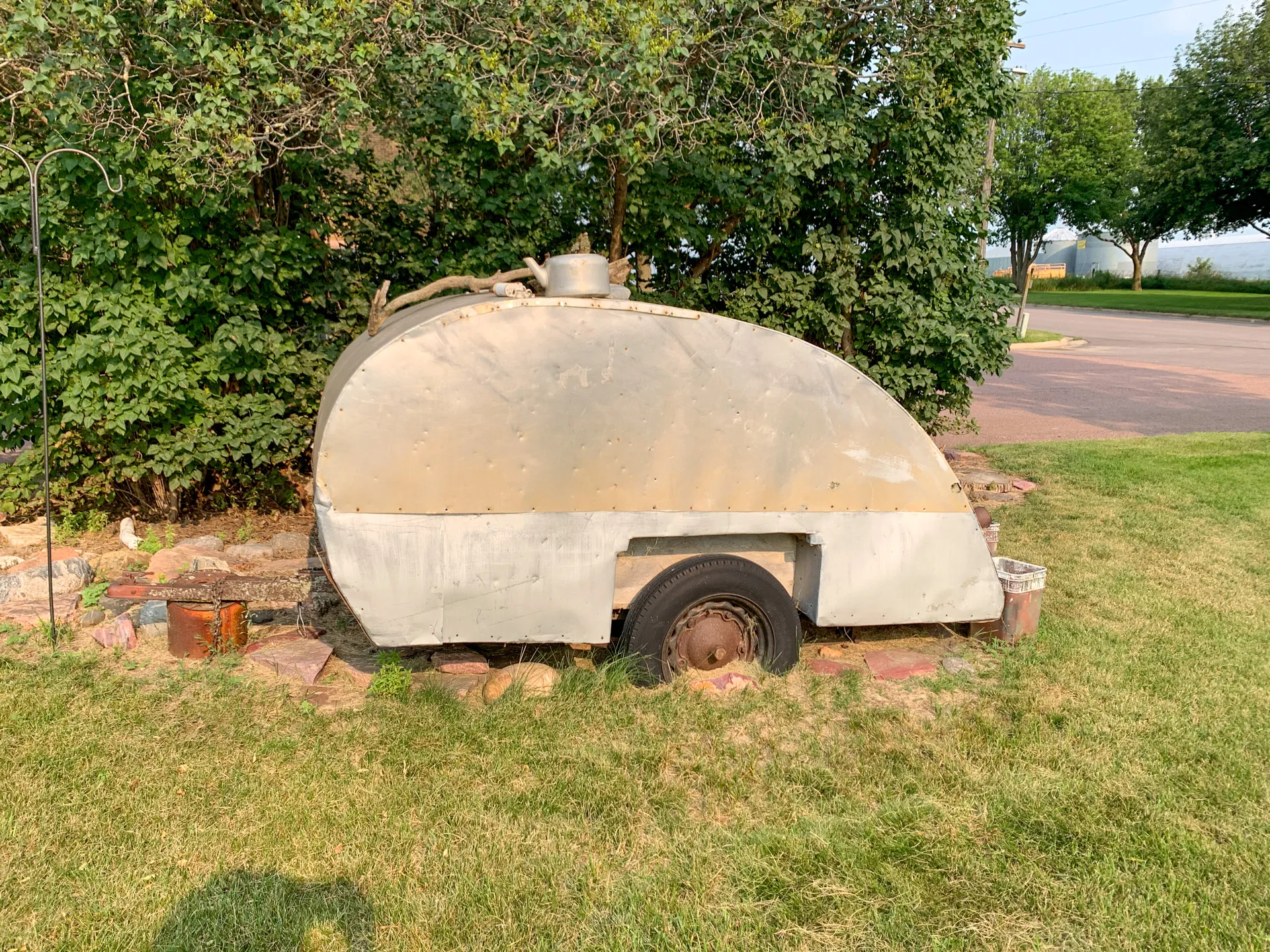 An old, weathered, gray trailer with a rounded top is positioned on a grass lawn near a line of trees near DeBates Memorial Park in Jasper, MN, surrounded by rocks and small plants.