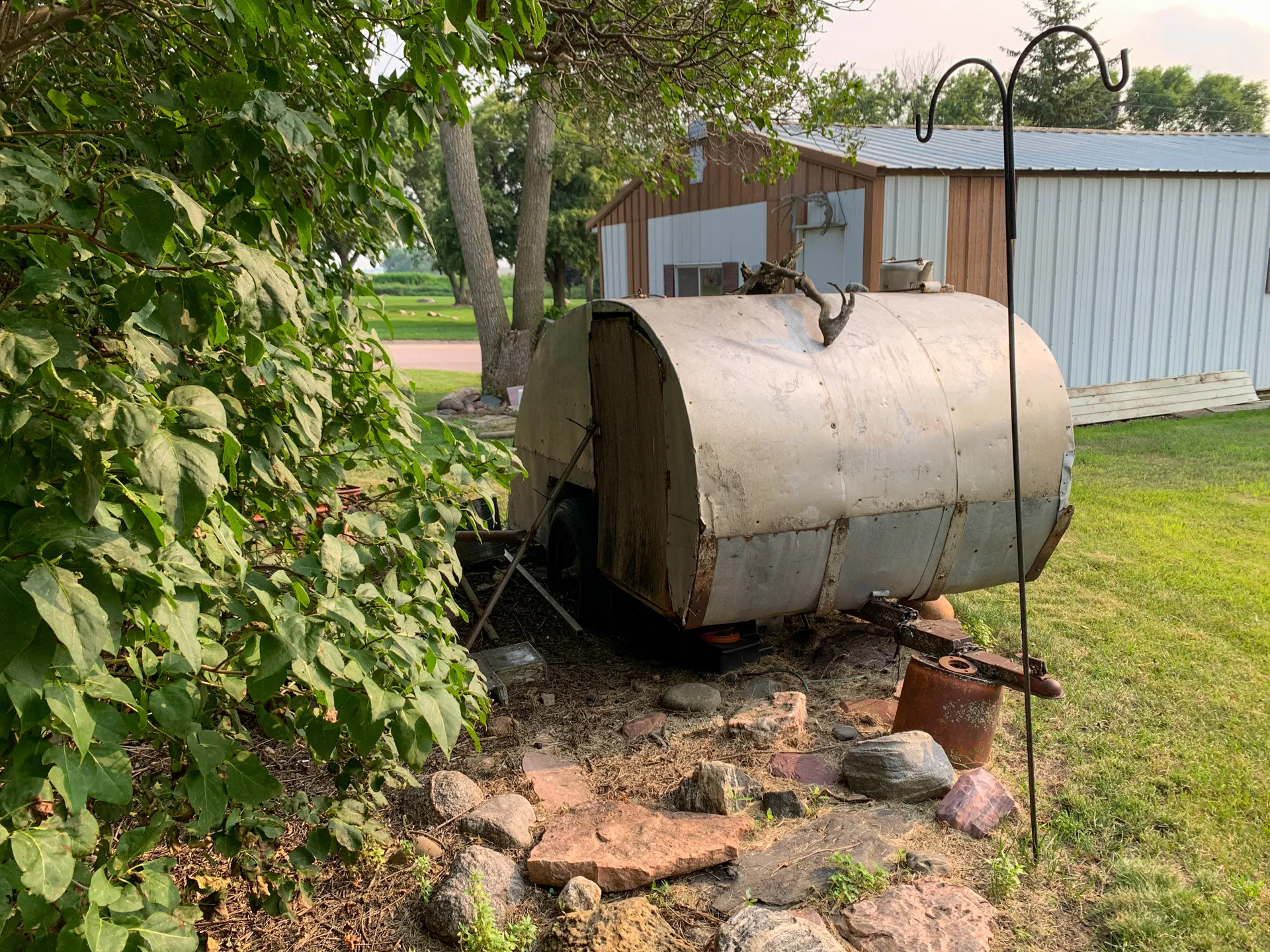 A rundown, rusted trailer near a small wooden building in a grassy yard. Trees and bushes surround the trailer, while stones with a metal pole and rusting pot lie in the foreground, creating a picturesque yet eerie scene.