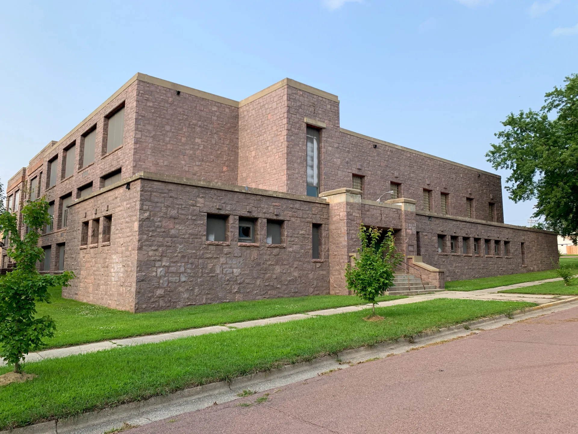 Located in Jasper, the large rectangular brick building was once a school and boasts a minimalistic design with small windows and a slightly elevated entrance with steps. It sits gracefully on a green lawn beside a paved street adjacent to the serene DeBates Memorial Park in MN.