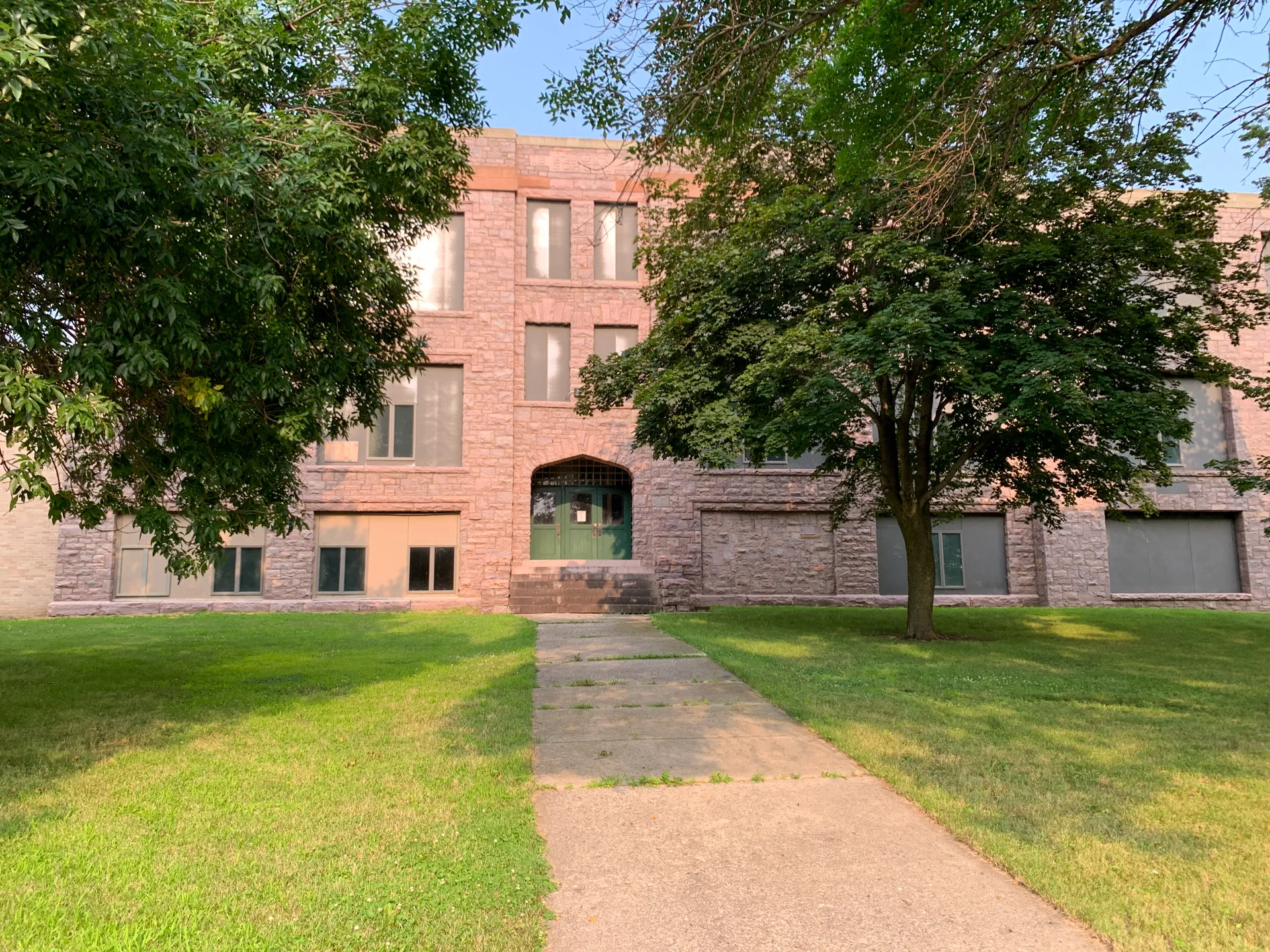 A three-story brick school with multiple windows stands proudly in DeBates Memorial Park, MN. Surrounded by green grass and trees, and with a paved walkway leading to its entrance, it's a serene spot complemented by the town's signature jasper.