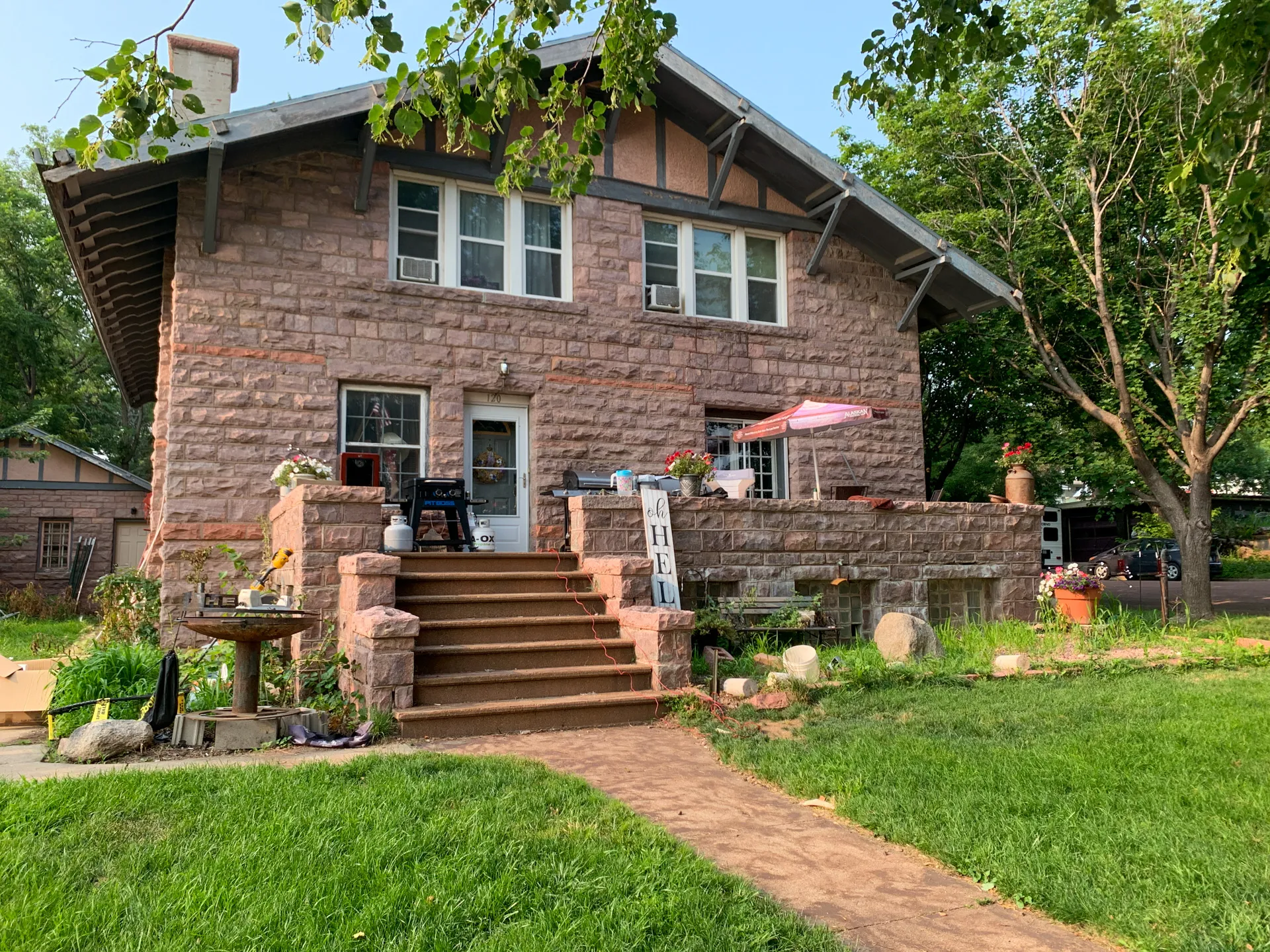 A two-story stone house in Jasper, MN, with a small front porch and steps leading to the entrance. Air conditioners sit in the windows, while a well-maintained garden surrounds the property with greenery and plants. Just a short walk away is DeBates Memorial Park.