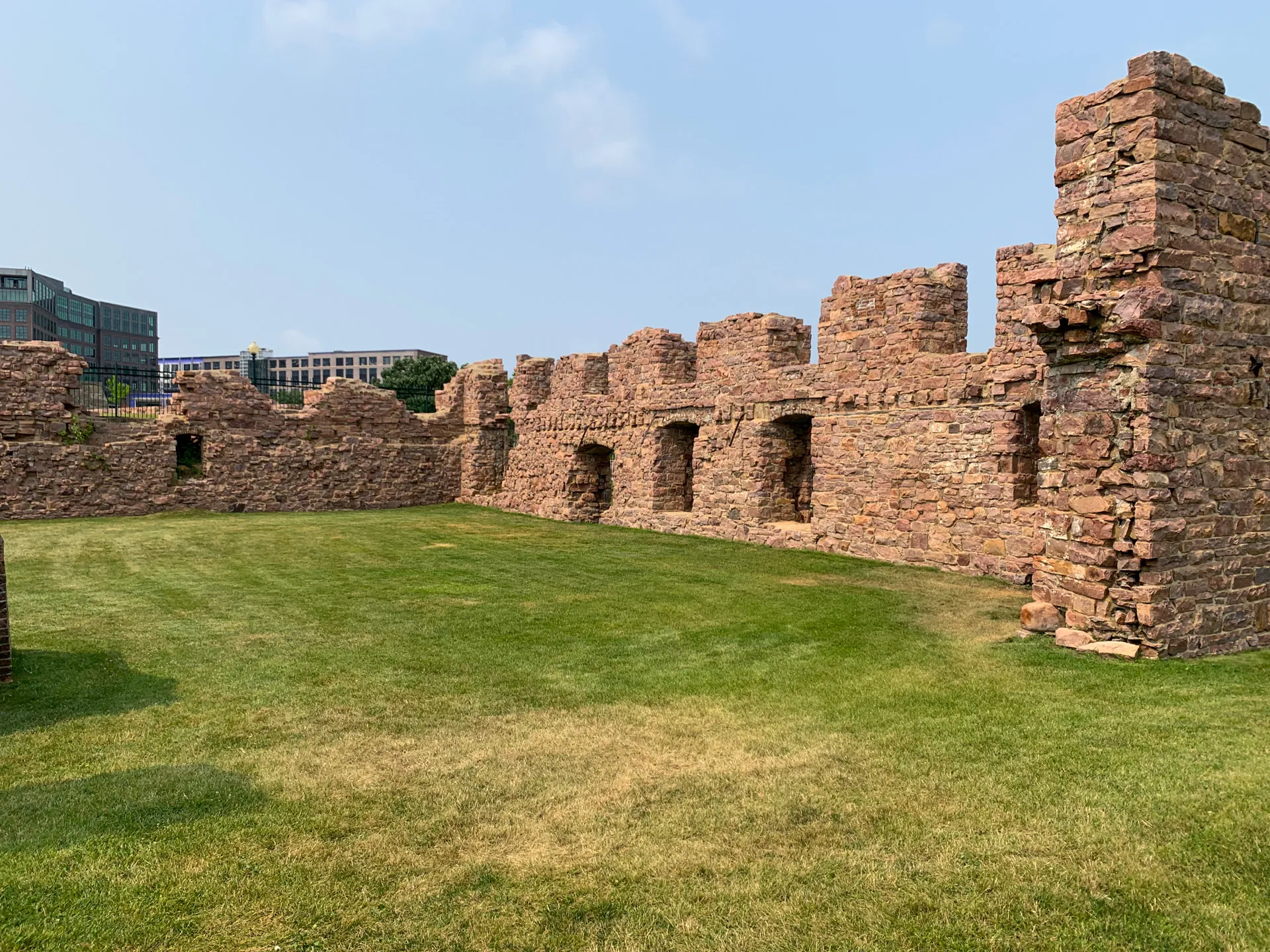 The stone ruins of an old building, with partial walls and turrets, stand in a grassy area at Falls Park in Sioux Falls, SD, with modern buildings rising in the background.