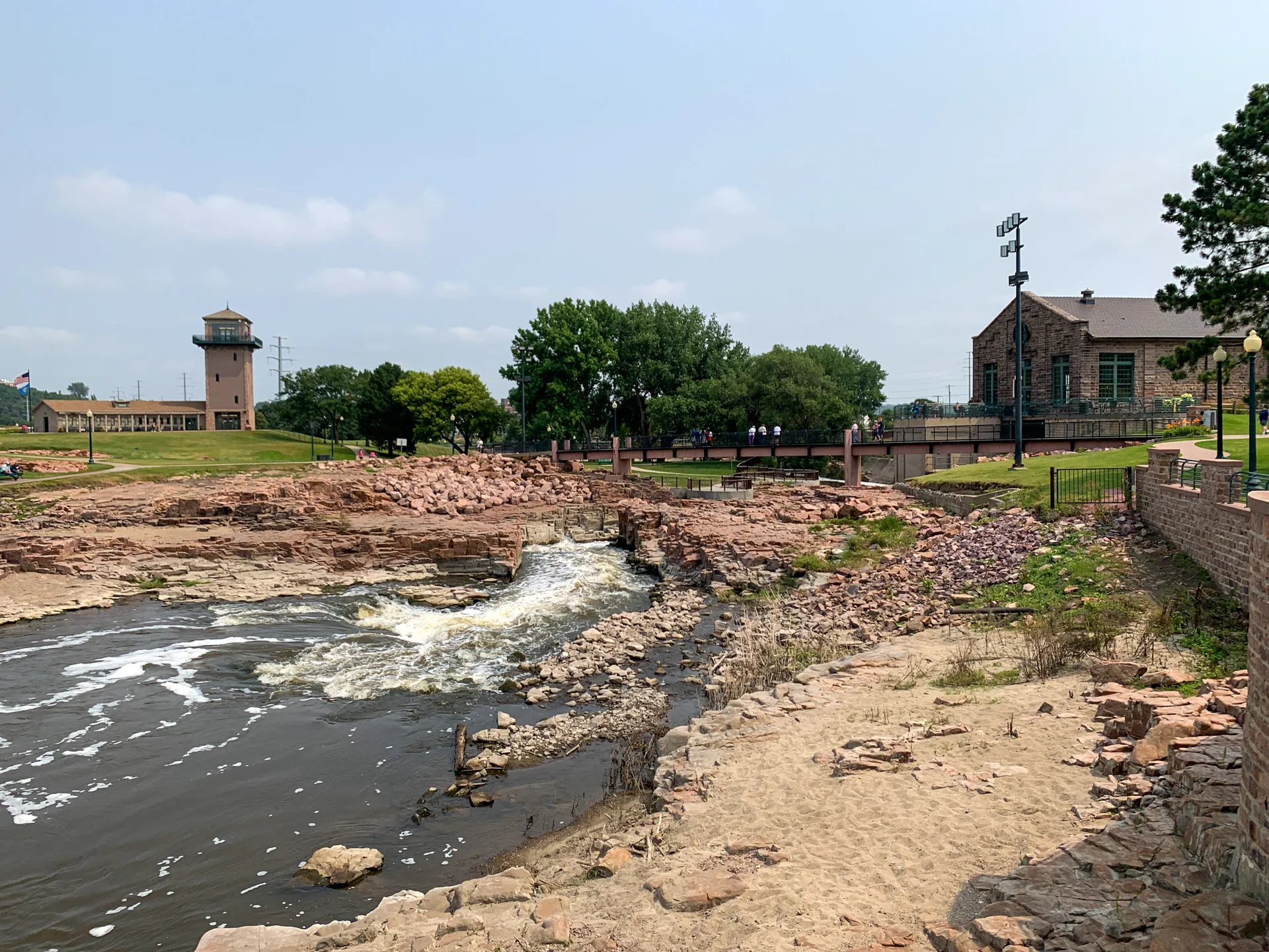Rocky riverbank with flowing water in a park setting, surrounded by trees and buildings in the background under a clear sky at Falls Park, Sioux Falls, SD.