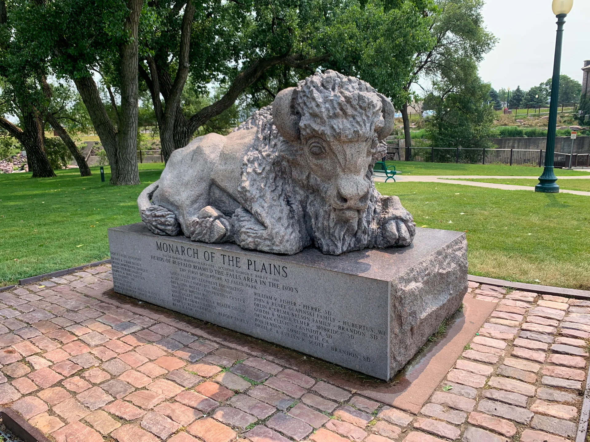 The "Monarch of the Plains" statue, depicting a majestic bison on a stone pedestal, graces Falls Park in Sioux Falls, SD, with trees and a lamp post enhancing its outdoor setting.