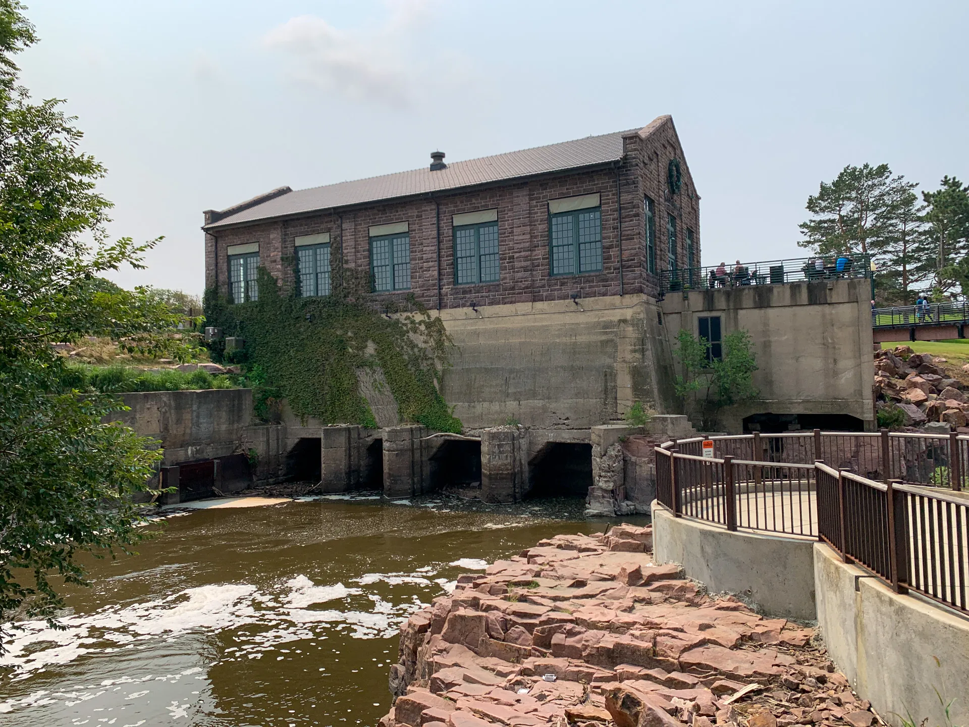 A brick and concrete industrial building is located by a flowing river in Falls Park, with several large openings at the water's edge, surrounded by trees and a stone pathway in Sioux Falls.