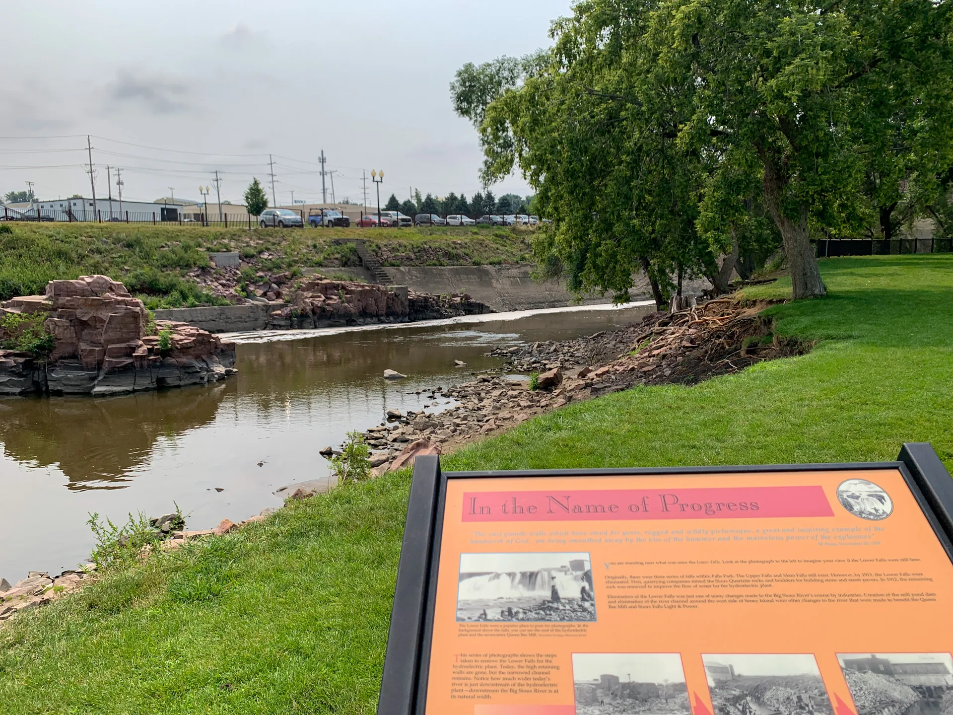A river bordered by grassy land and trees with an informational plaque in the foreground titled "In the Name of Progress." In the background, vehicles and buildings visible beyond the riverbank hint at the bustling growth of Sioux Falls, SD.