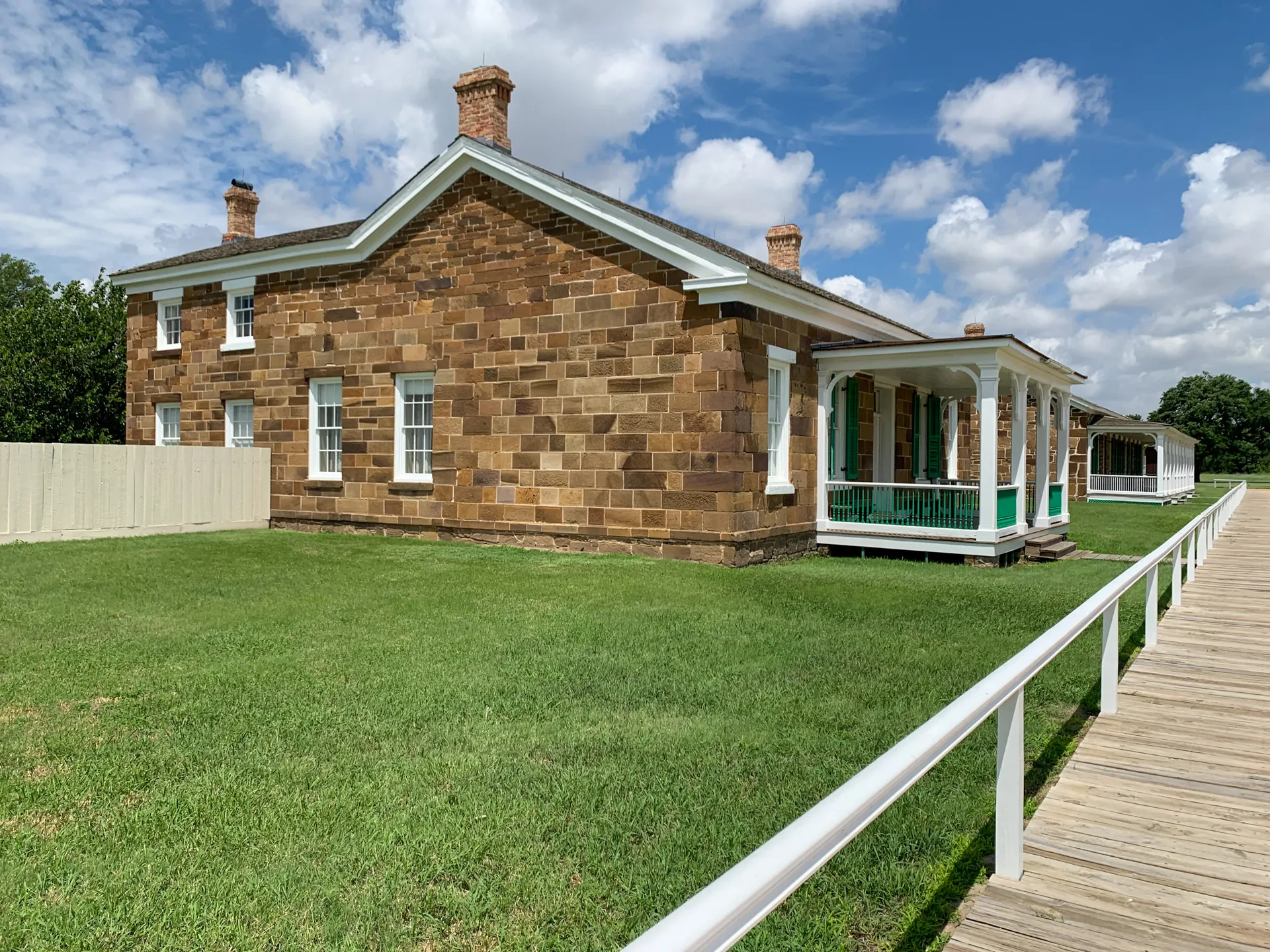 A historic stone building with a green porch, white trim, and a boardwalk leading to the entrance, set against a backdrop of a grassy lawn and partly cloudy sky.