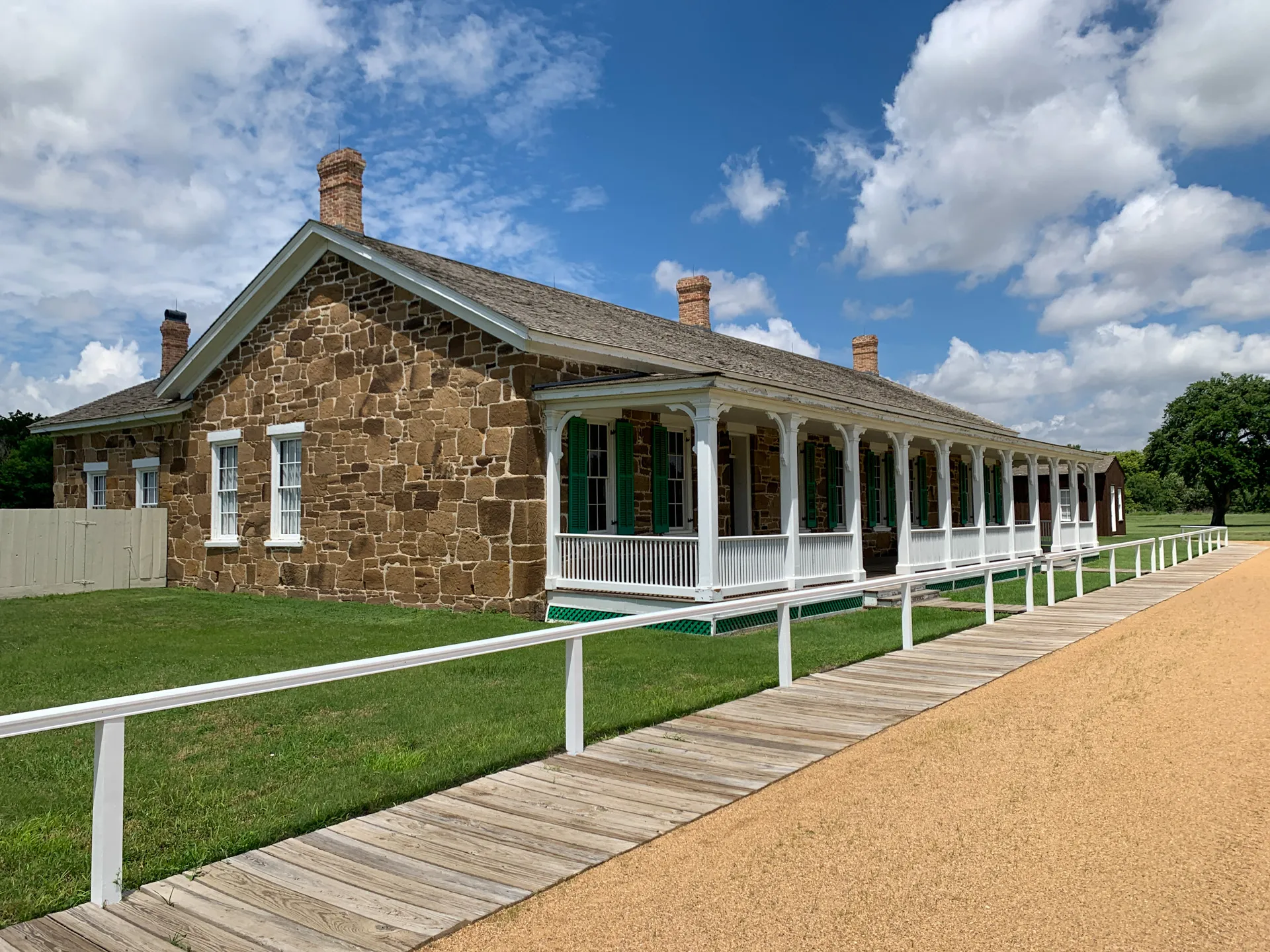 A long, single-story stone brick building with a wooden porch and green shutters sits under a partly cloudy sky. A wooden walkway and gravel path run alongside the building.