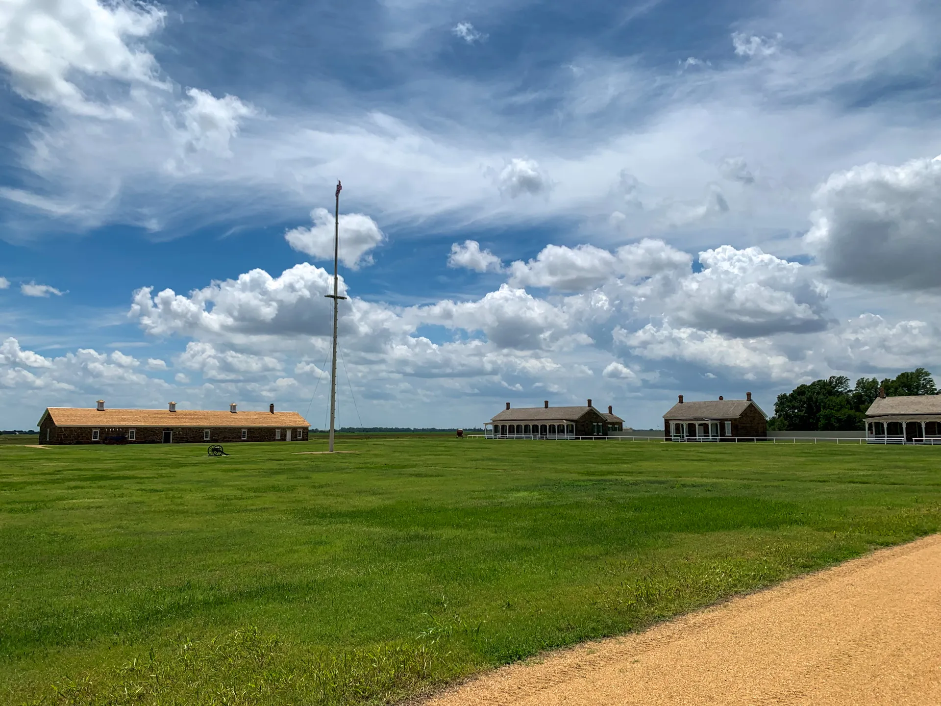 A grassy field with several buildings in the background. A flagpole stands in the middle of the field. A dirt path runs along the right side under a partly cloudy sky.
