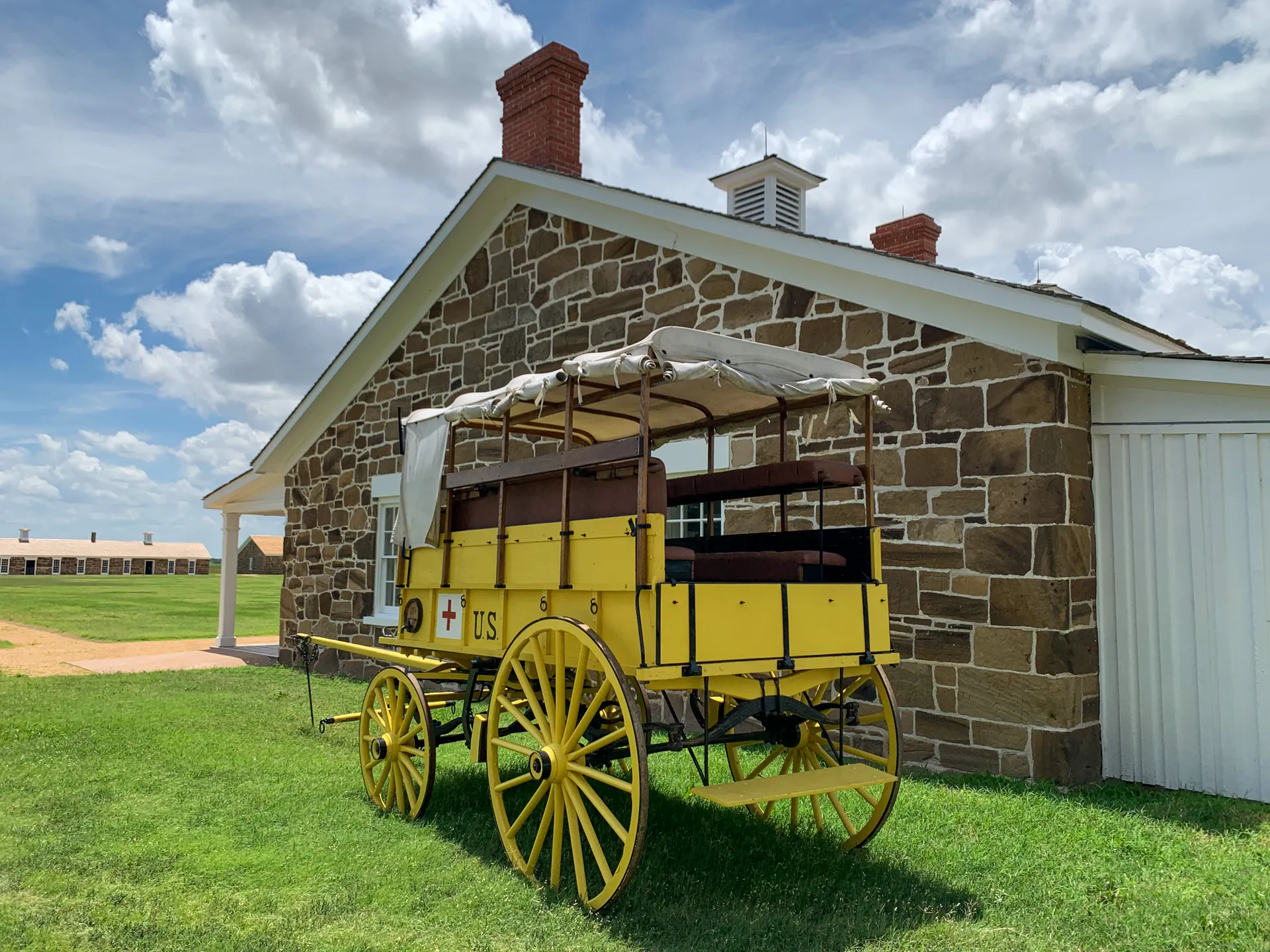 A yellow horse-drawn wagon with a white cover, labeled "US" and marked with a red cross, is parked in front of a stone building on a grassy area under a partly cloudy sky.