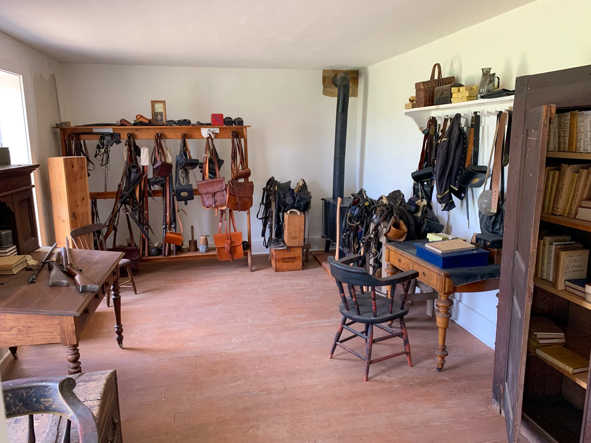 A neatly arranged room with shelves of books and bags, a desk with papers, and various leather goods hanging. There's a wood stove in the corner and a wooden chair near a table.