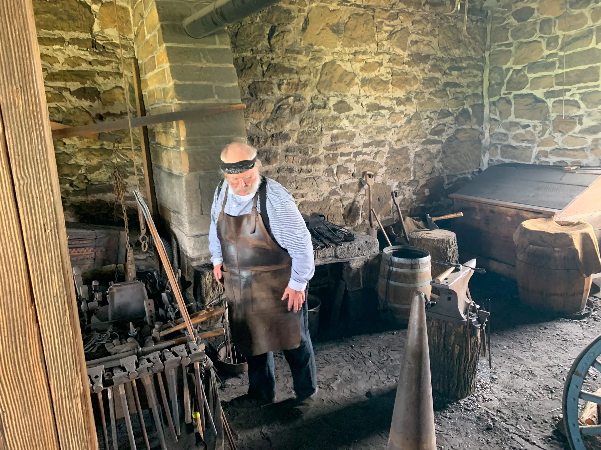 A blacksmith wearing a protective face shield and apron works in a rustic stone forge with an anvil, barrels, and various tools surrounding him.