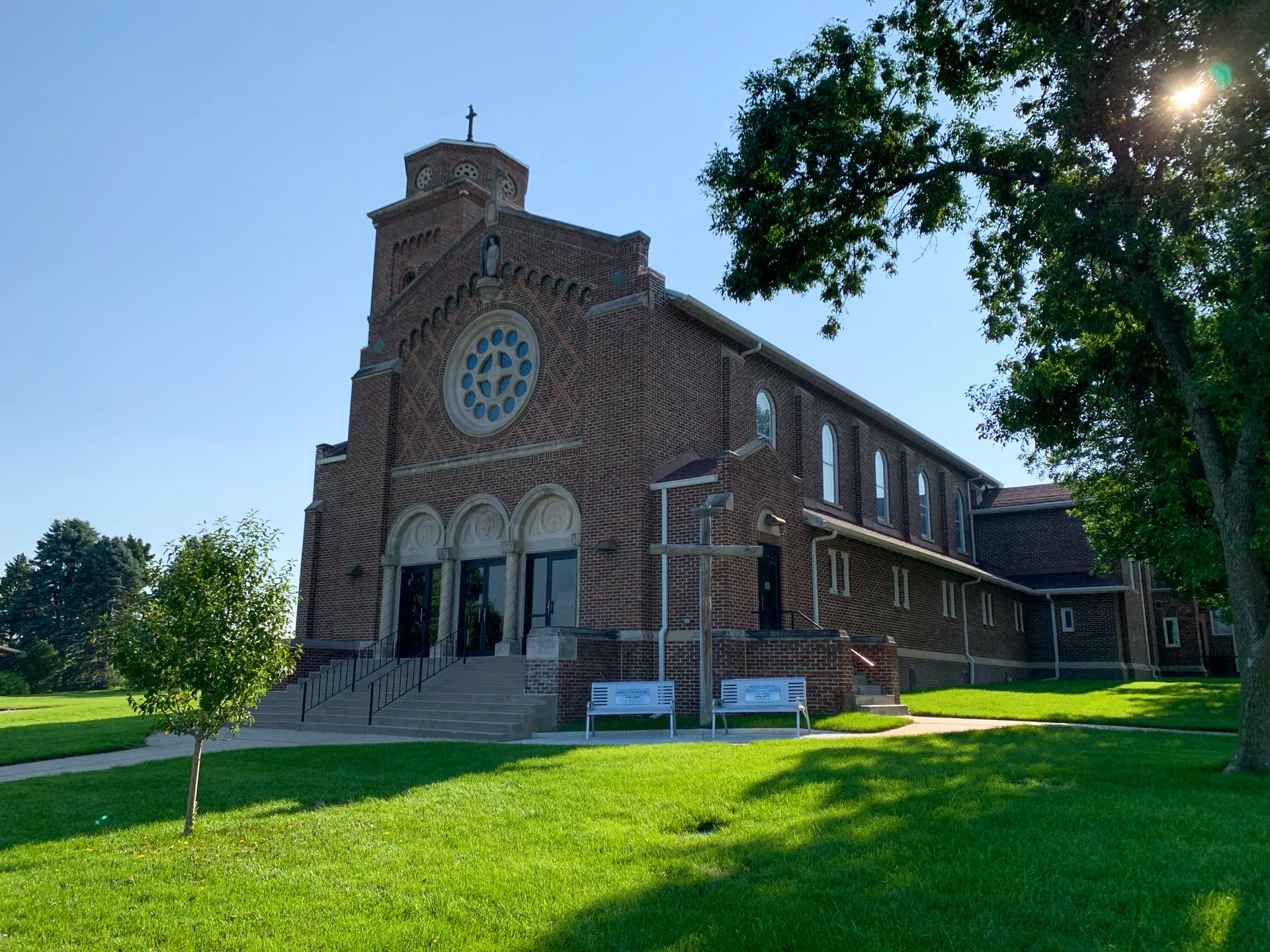 A red-brick building, St Joseph Catholic Church, with arched entryways, a rose window, and a cross on top, surrounded by green grass and trees on a sunny day.