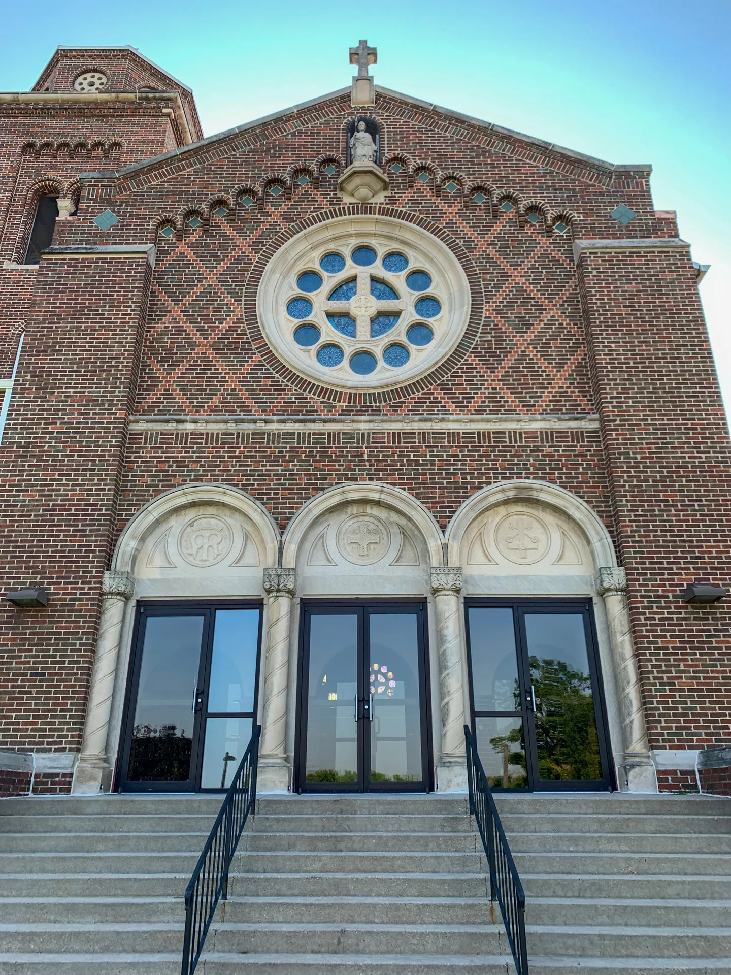 The front facade of St Joseph Catholic Church features a large circular stained glass window, three arched entrances with glass doors, and a staircase leading up to the doors.