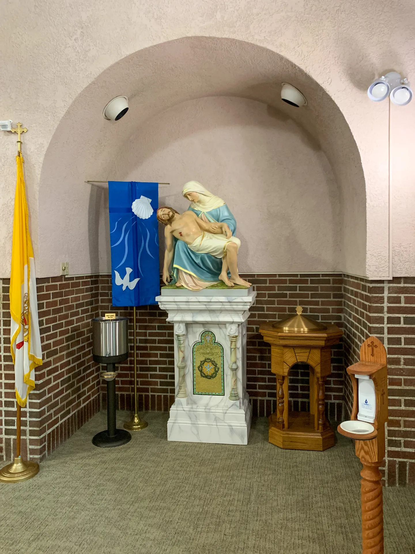 A religious statue depicting Mary holding Jesus is displayed on a pedestal, flanked by flags, a candle holder, and a lectern in a room with arched and beige walls at St. Joseph Catholic Church.