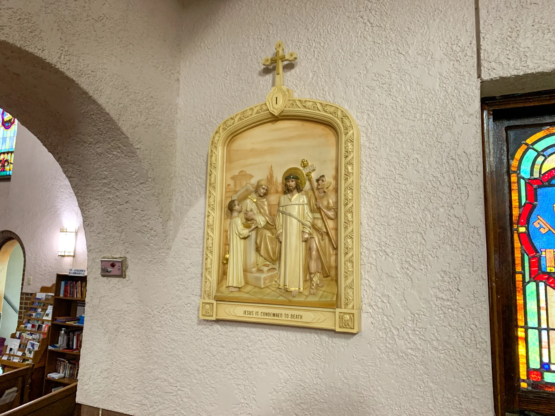 A relief sculpture on a Catholic Church wall depicts a biblical scene titled "Jesus is Condemned to Death." A crucifix is mounted above the artwork.