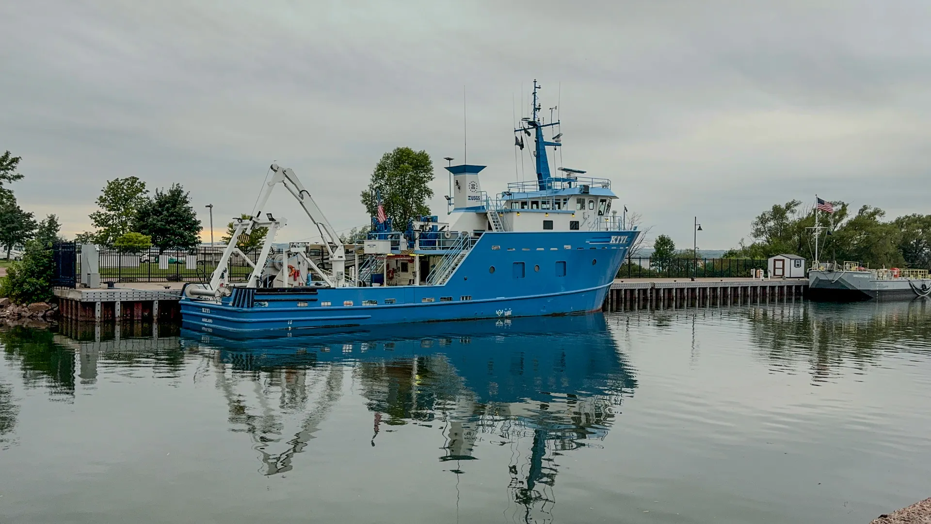 A blue research vessel is docked at a pier on a calm, overcast day in Kreher RV Park, Ashland, Wisconsin; reflections of the boat and surroundings are visible on the water.