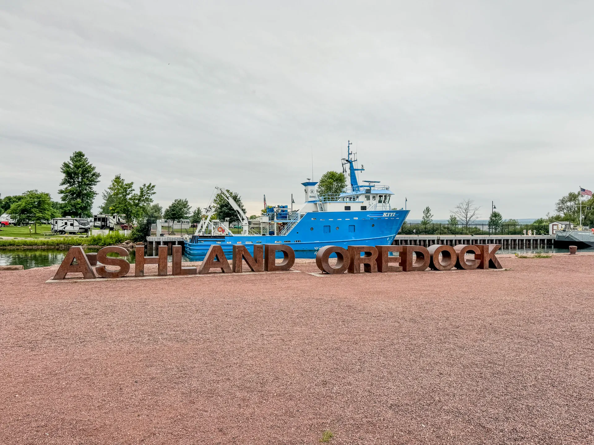 A landscape featuring a large sign reading "Ashland Oredock," with a blue ship docked nearby on a cloudy day, set against the serene backdrop of Ashland, Wisconsin, near the Kreher RV Park.