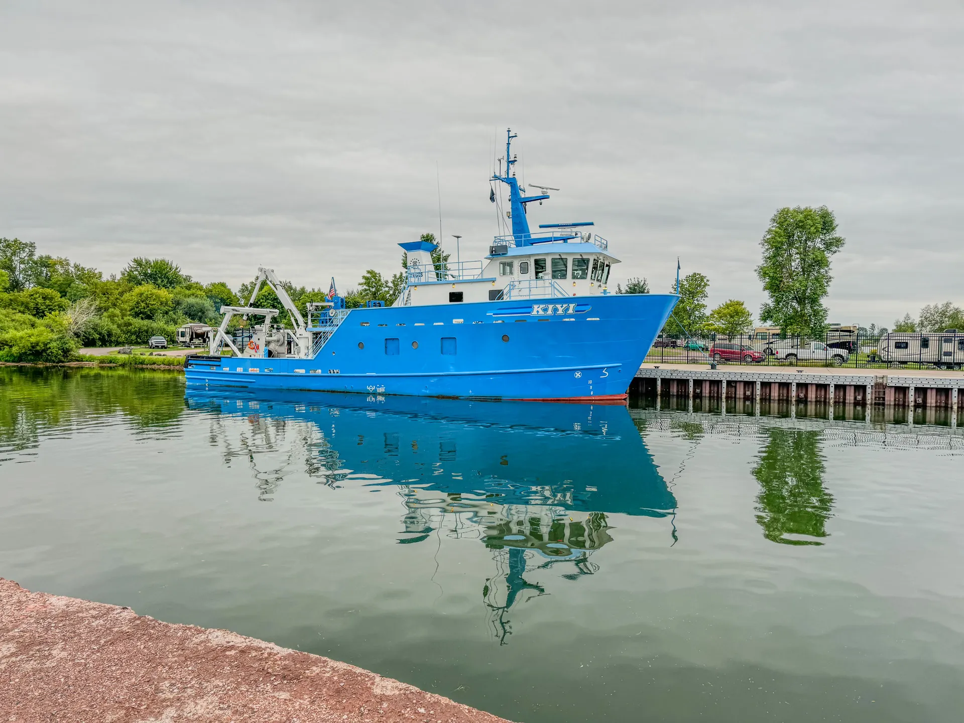 A large blue research vessel named "Kiyi" is docked in the calm harbor at Kreher RV Park in Ashland, Wisconsin, its reflection shimmering on the water. Trees and parked cars line the background.