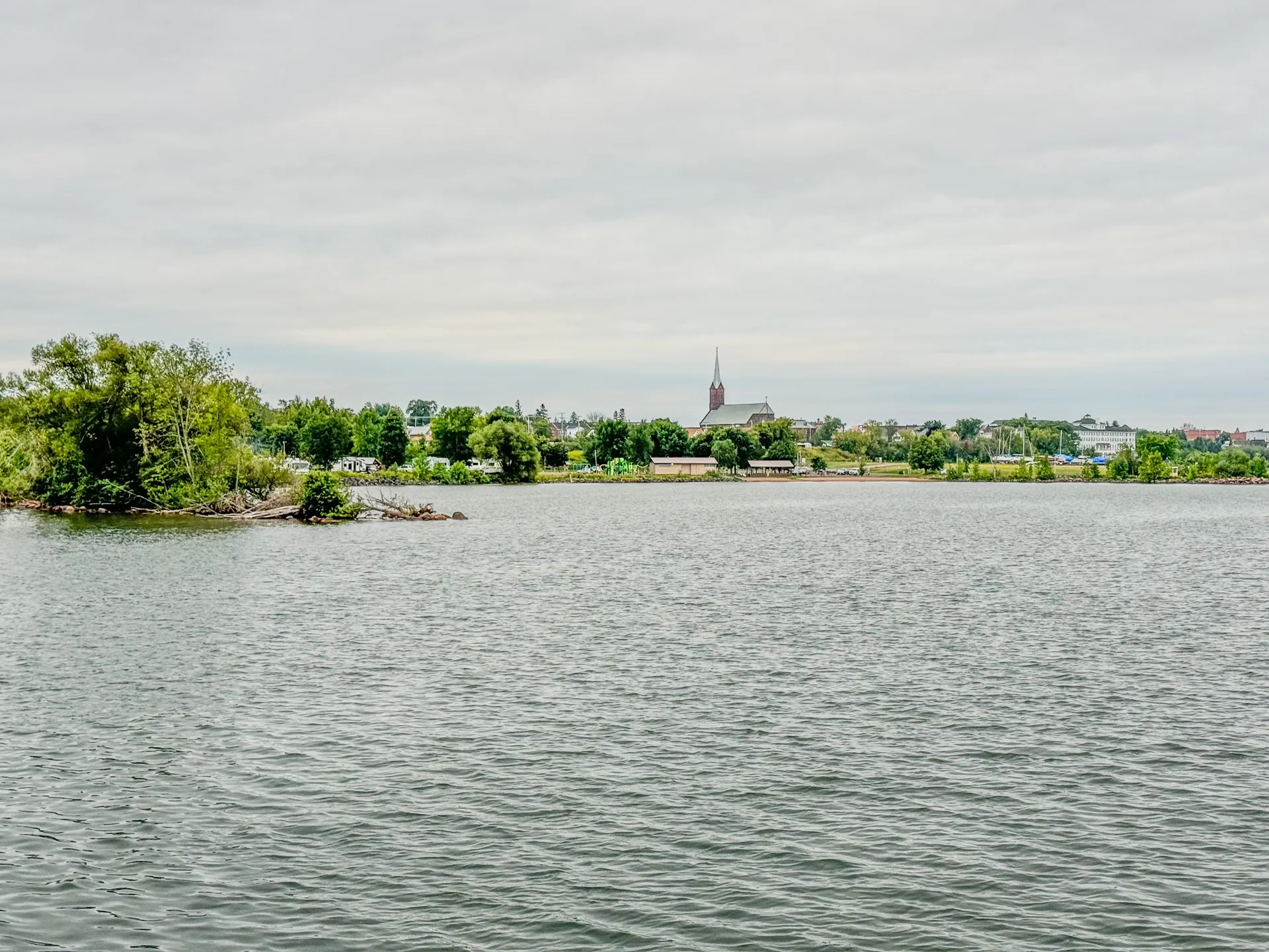 A calm body of water with a distant shoreline featuring trees and buildings, including a church with a steeple under a cloudy sky, embodies the serene charm of Ashland, Wisconsin near Kreher RV Park.