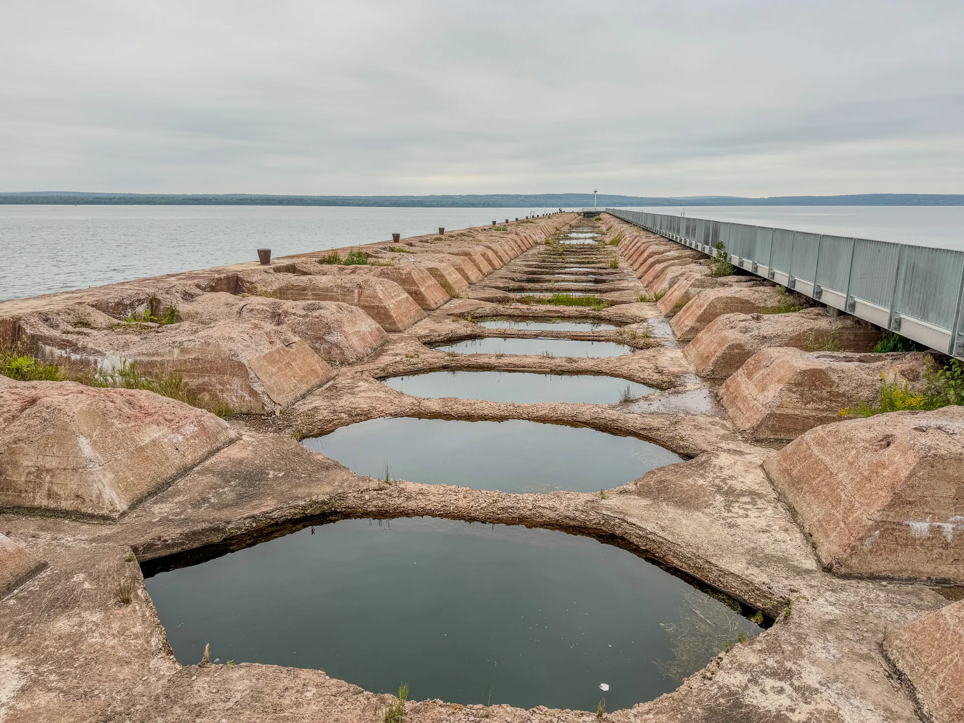 A stone pier extends into a body of water at Ashland's Kreher RV Park in Wisconsin, with large hexagonal rocks partially submerged. A metal railing lines the right side of the pier under an overcast sky.