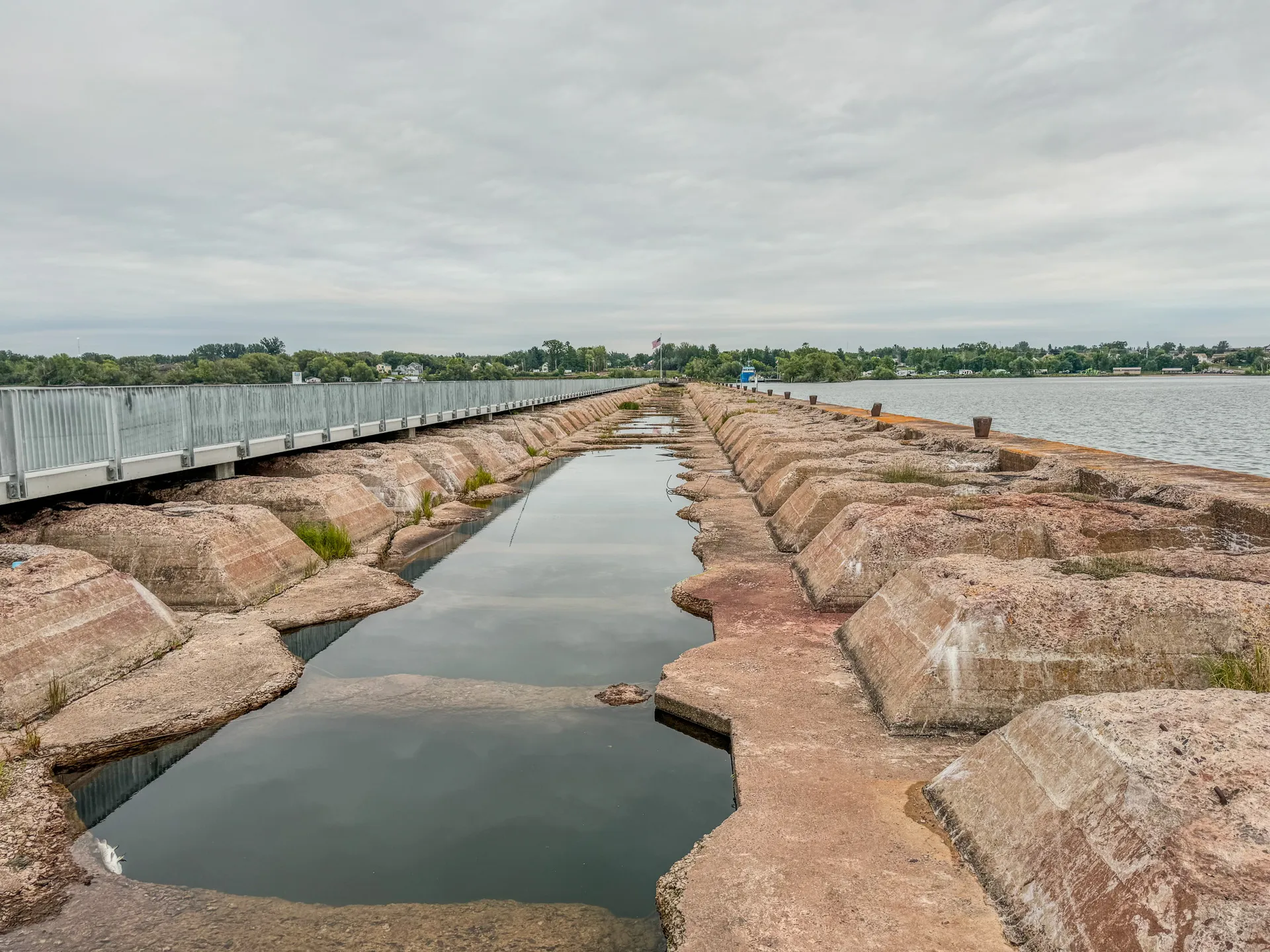 The concrete pier, featuring a series of rectangular blocks and pools of water, extends into a body of water under a cloudy sky at Ashland's Kreher RV Park in Wisconsin. A metal fence runs along the side, with trees visible in the background.