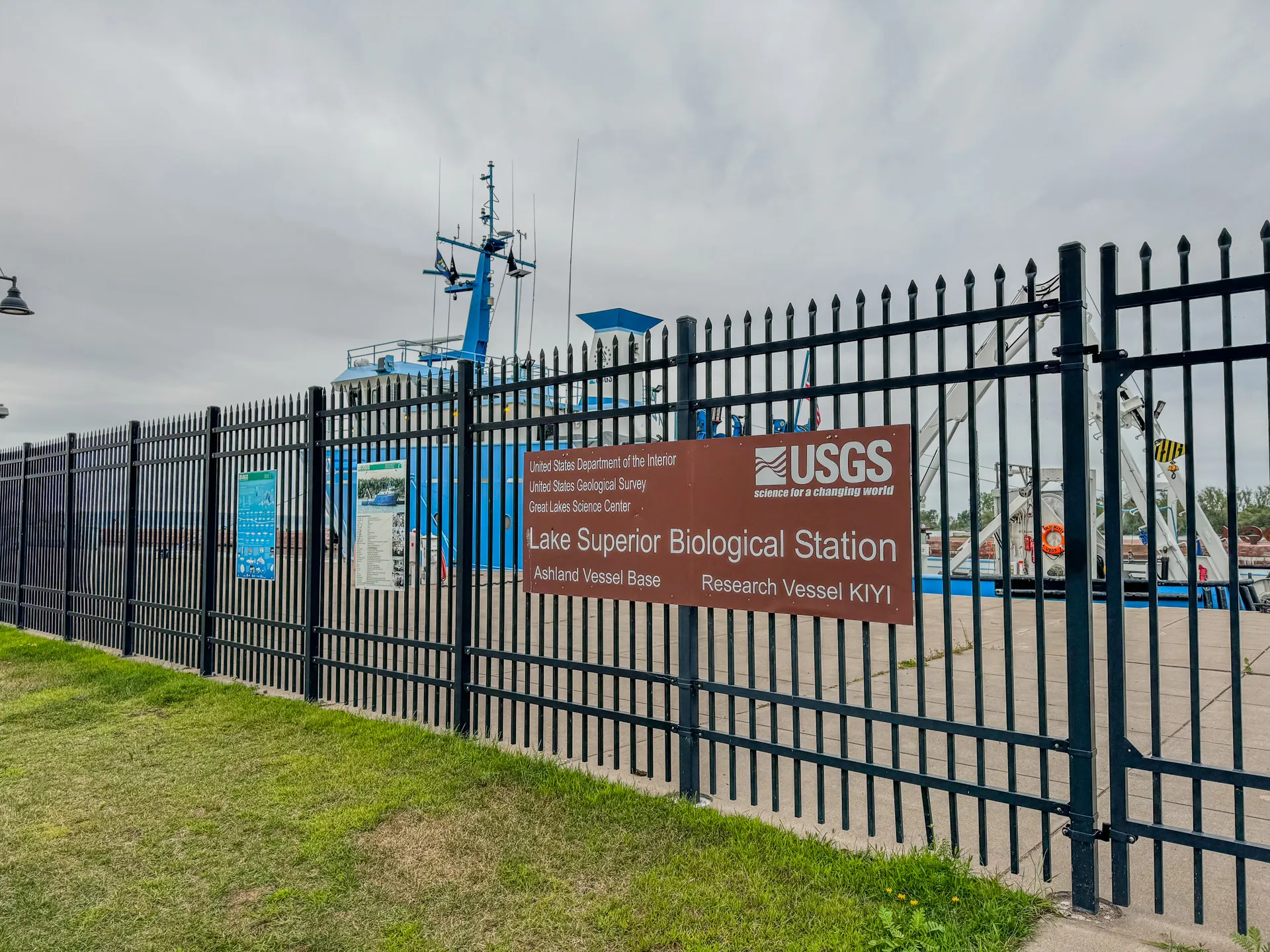 A fenced area with a sign for the USGS Lake Superior Biological Station in Ashland, Wisconsin, is situated in front of a docked research vessel named KIYI, near the serene Kreher RV Park.