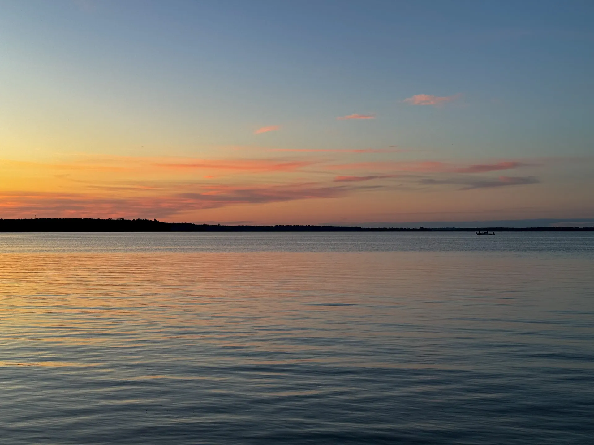 A calm body of water reflecting hues of pink, orange, and blue from a sunset at South Pike Bay Campground in Chippewa National Forest, with a silhouetted shoreline and a boat in the distance.
