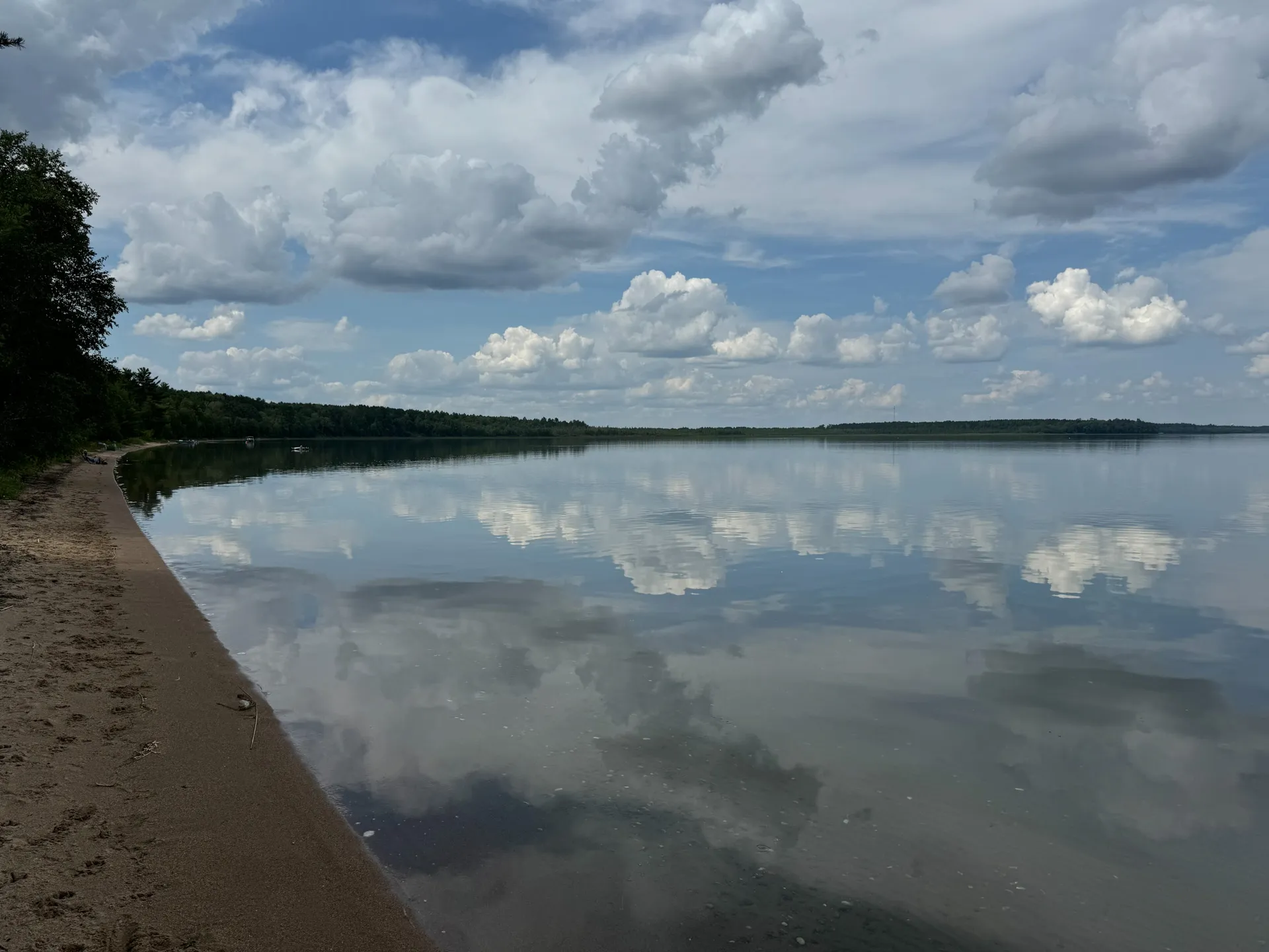 A calm lake reflects a blue sky with scattered clouds, with the sandy shore visible in the foreground and a dense tree line in the distance at South Pike Bay Campground, nestled within Chippewa National Forest.