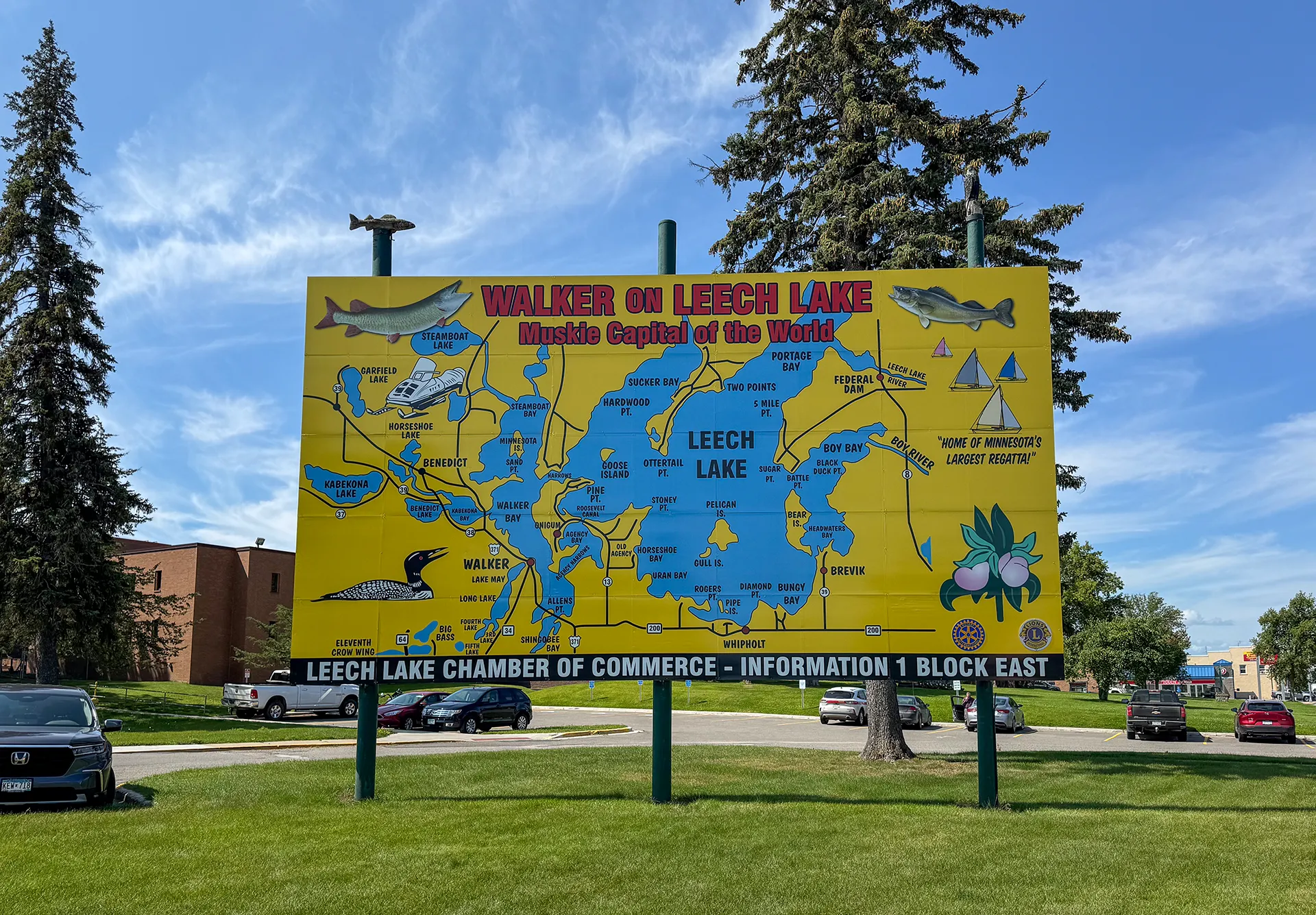 Large outdoor billboard with a colorful map of Leech Lake, highlighting Walker, MN as the "Musky Capital of the World." The sign, near Downtown Walker, is surrounded by trees and a grassy area with parked cars nearby.