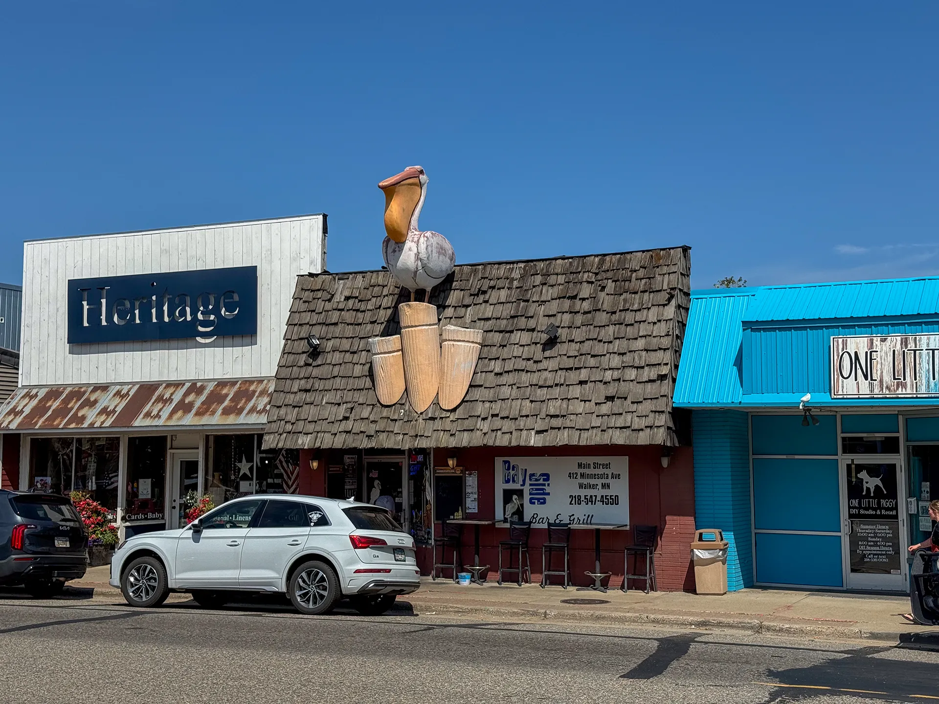 A storefront with a large pelican statue on the roof sits between two neighboring buildings in downtown Walker, MN. Three cars are parked on the street in front of the shops, adding to the local charm reminiscent of a cozy lighthouse town.