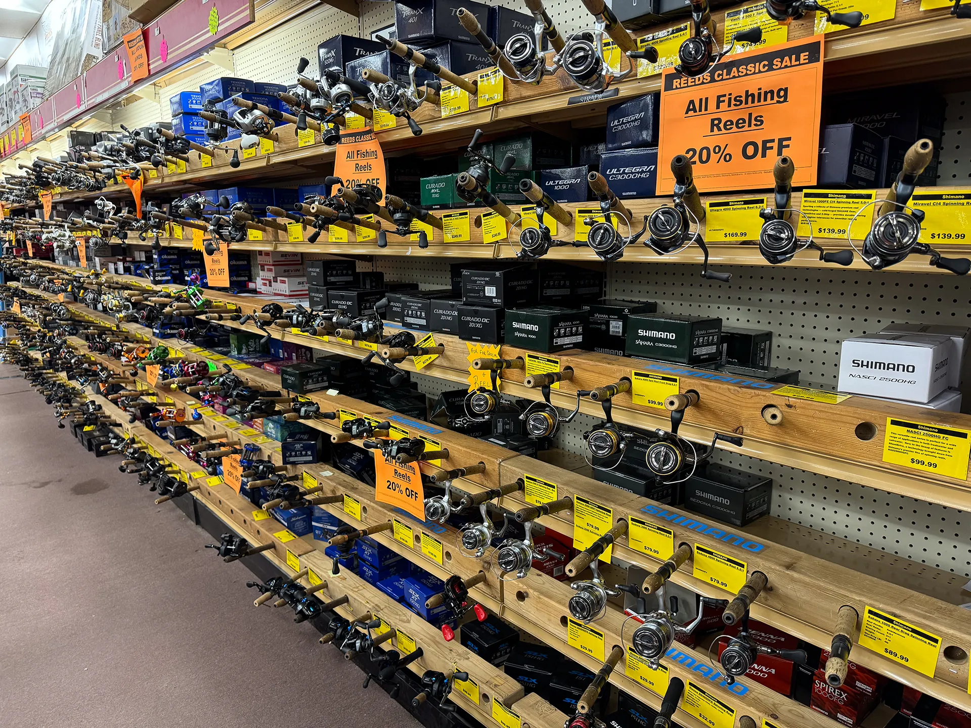 In a store aisle with shelves displaying various fishing reels and equipment, orange signs above indicate a 20% discount sale on all fishing reels. This downtown Walker, MN location is perfect for any angler's needs.