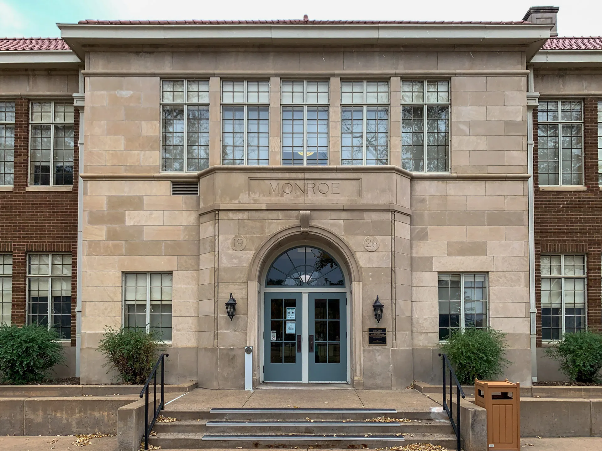 Exterior of a historic building with "Monroe" engraved above the entrance. The structure features large rectangular windows, a central double door, and symmetrical design with light-colored stone facade contrasted by darker brick sections. Two lampposts flank the doorway, and bushes adorn the sides.