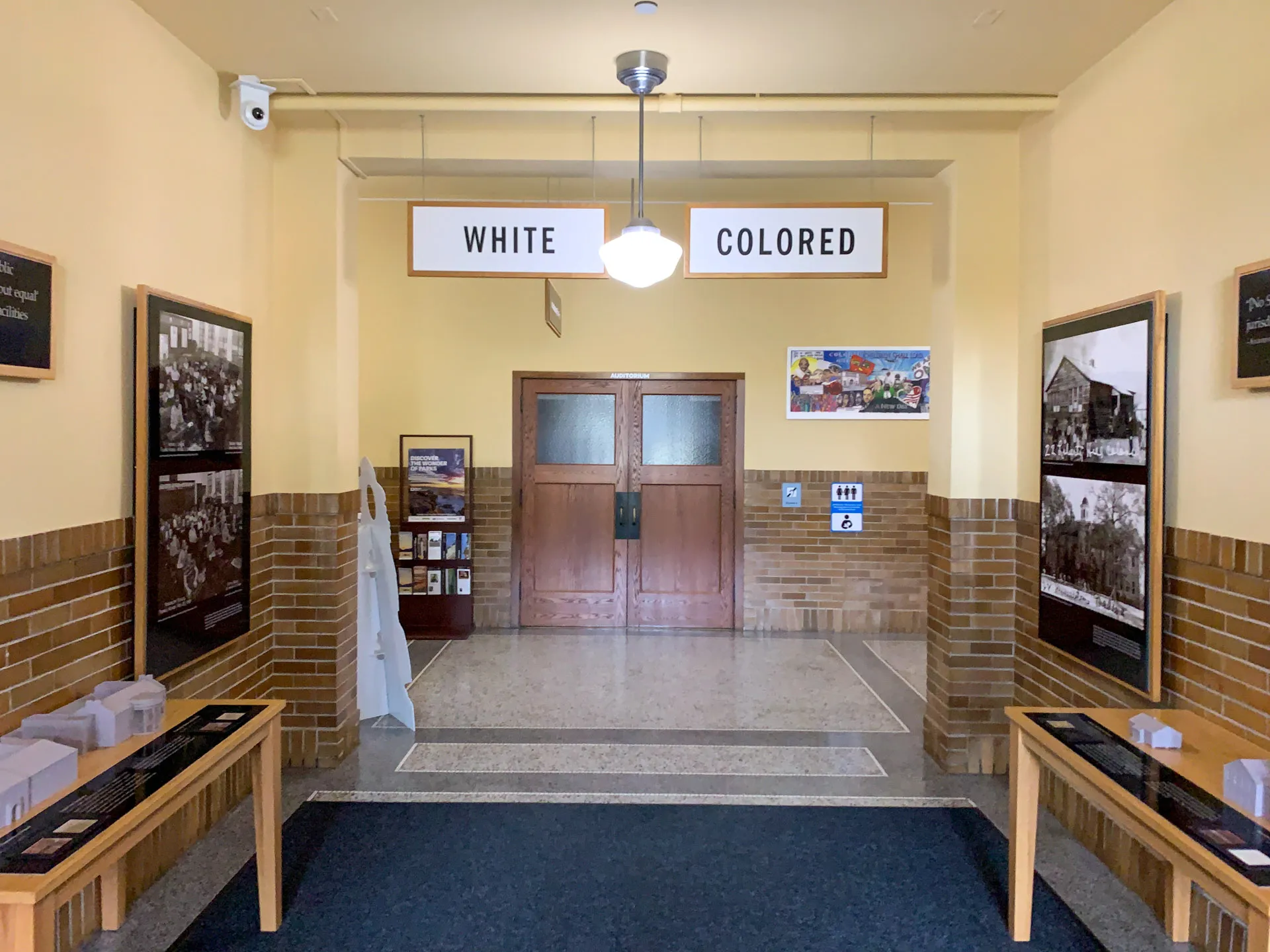A hallway with a vintage setting displays a historical exhibition. Signs reading "WHITE" and "COLORED" hang from the ceiling, representing racial segregation. The space has beige walls, wooden doors, and historical photos and information panels on both sides.
